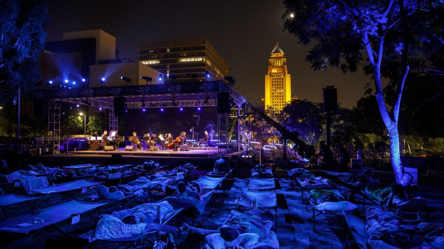 Outdoor nighttime concert with musicians on stage under purple lights, audience members lying on lounge chairs, city skyline with illuminated skyscraper in the background, and trees lit with blue lights.