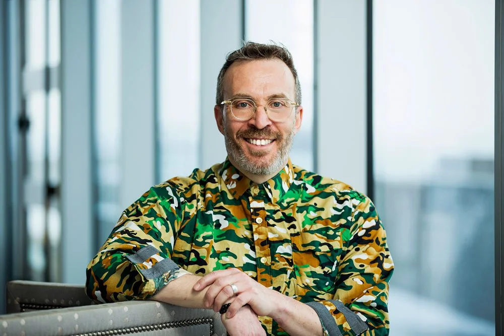A smiling man wearing glasses and a colorful camouflage-patterned shirt sits at a table in a modern office with glass walls.