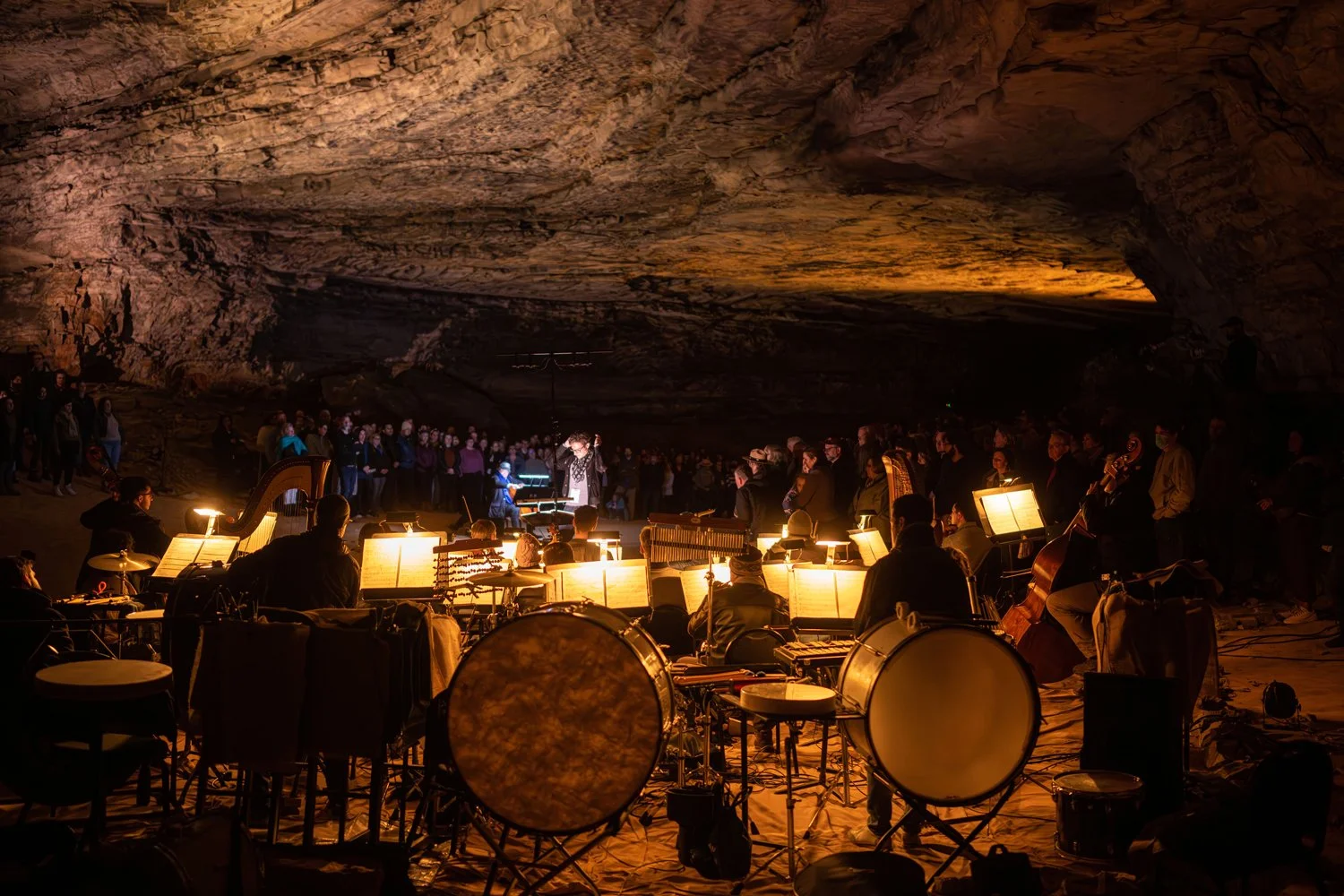 An indoor musical concert taking place inside a large cave with a rock ceiling. The stage is illuminated with warm lighting, featuring musicians with instruments and sheet music, and a large group of audience members watching.