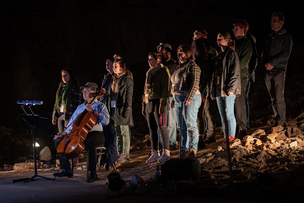 Group of people, including a cellist, performing on a stage with rocks, during a nighttime outdoor event.
