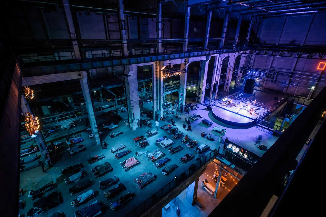 An aerial view of an indoor concert venue or event space with a stage, seating, and people lying on the floor.