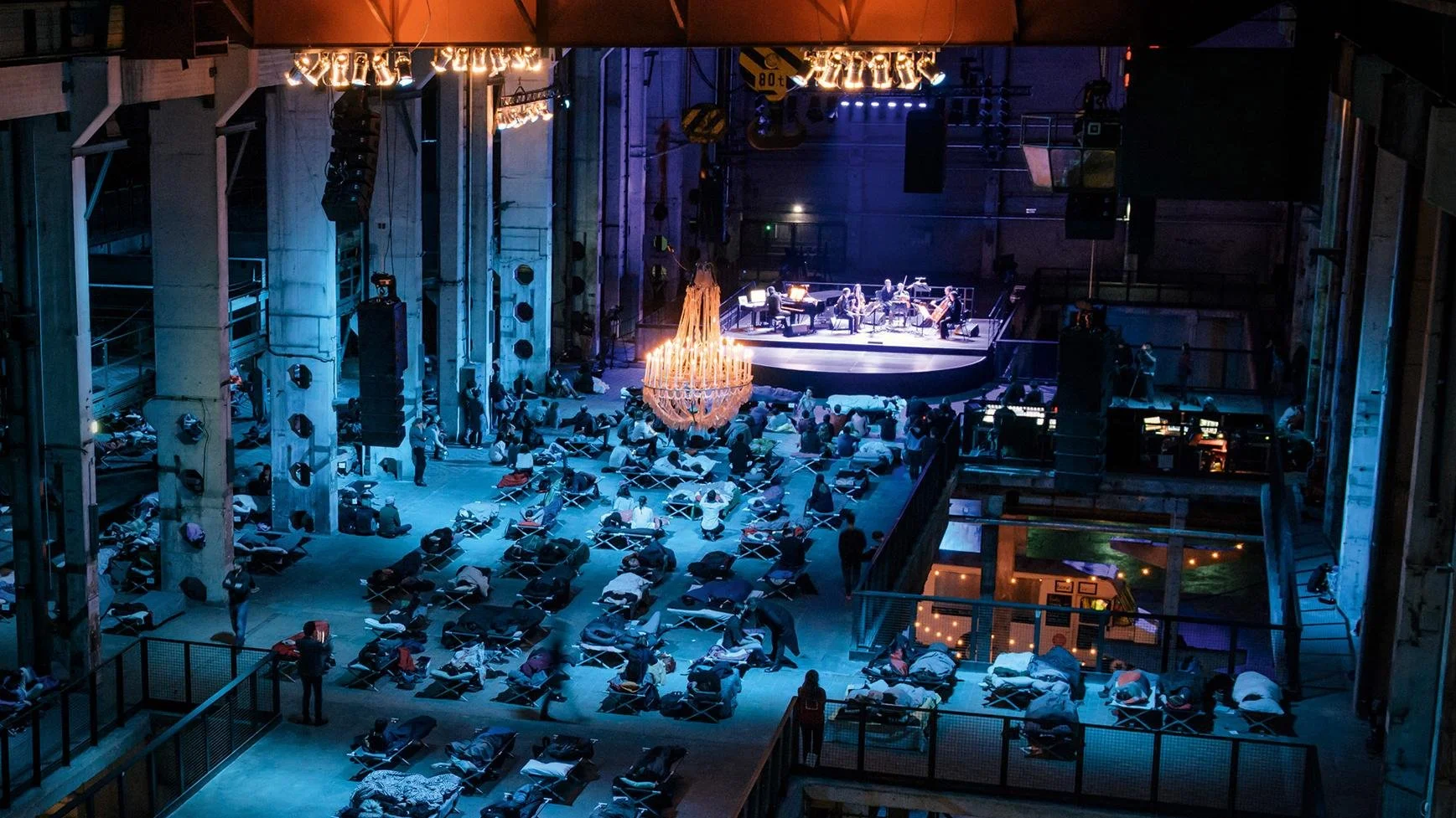 Indoor concert hall with a stage featuring a band, audience members seated or lying on cots, ambient lighting, and a chandelier hanging from the ceiling.