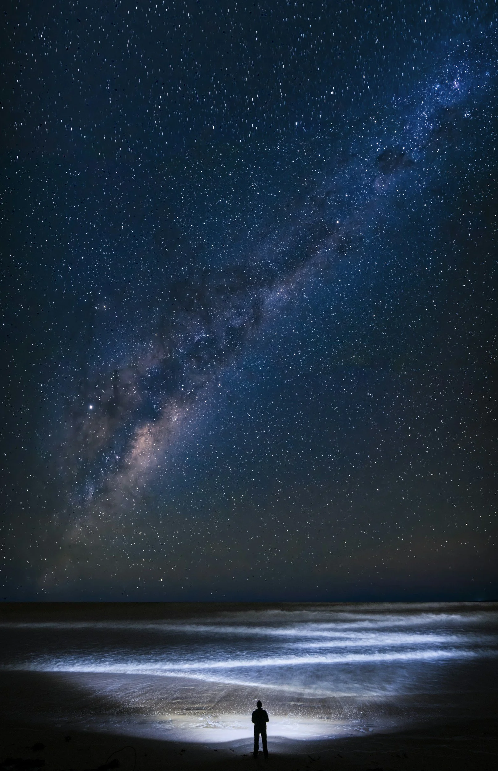 Concept image for a work in progress: seen from a distance a human gazes toward the vast ocean at night while the Milky Way Galaxy brims and overflows the sky with starlight.