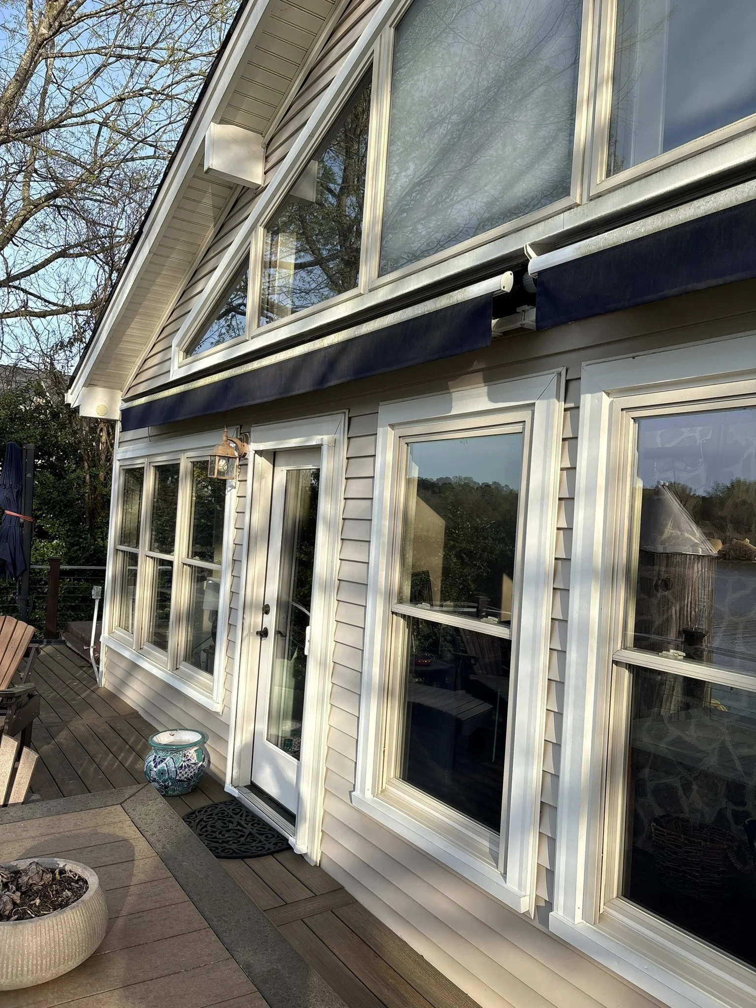 Rear exterior view of a house with large glass windows, a sliding glass door, an outdoor deck with wooden flooring, and a decorative ceramic pot on the deck. There are leafless trees reflected in the windows.