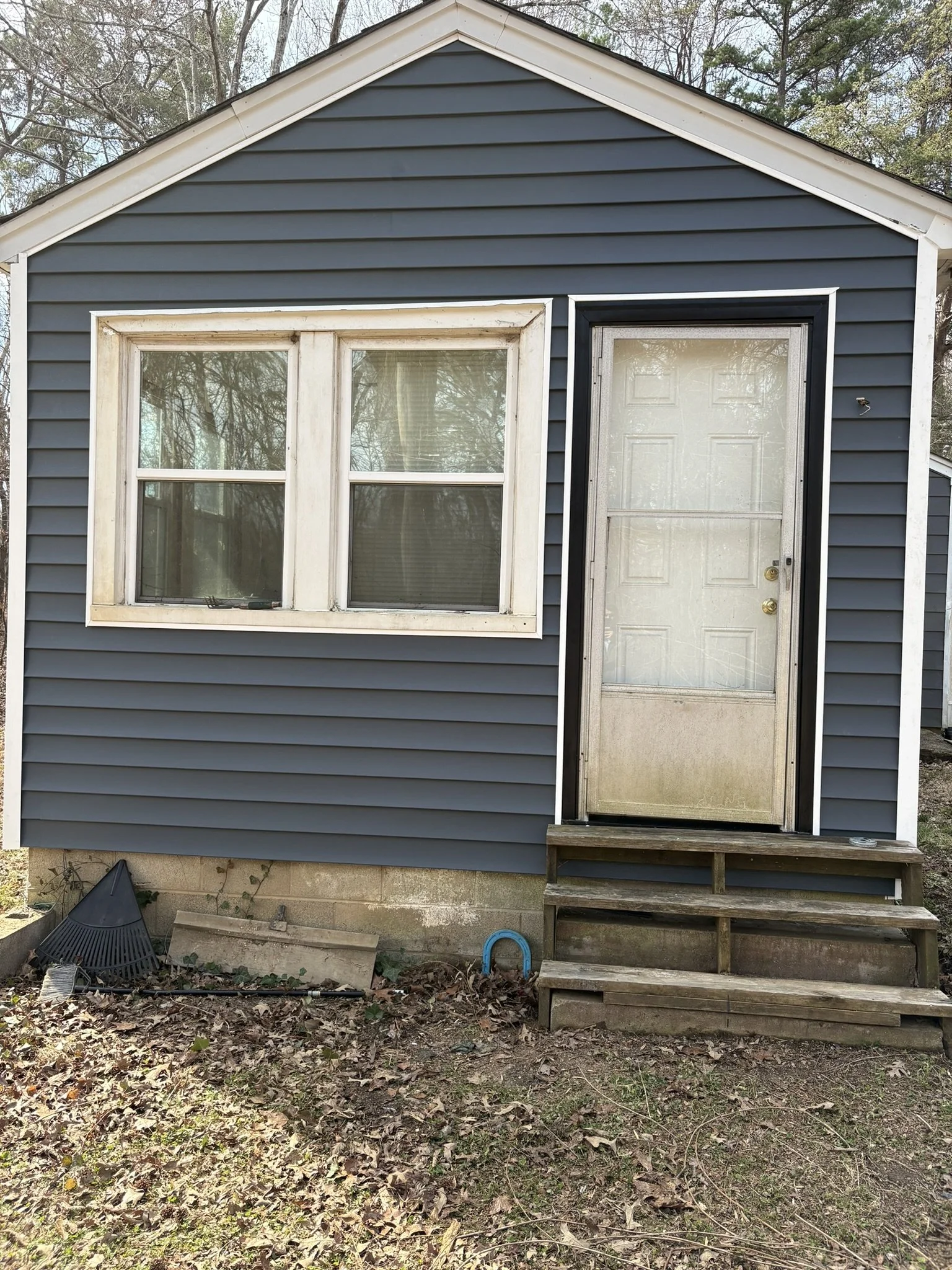 Backyard view showing the exterior of a blue house with beige trim, including a window and a door with a metal screen, along with wooden stairs leading to the door. The ground in front is covered with leaves and dirt, with some garden tools and a gar