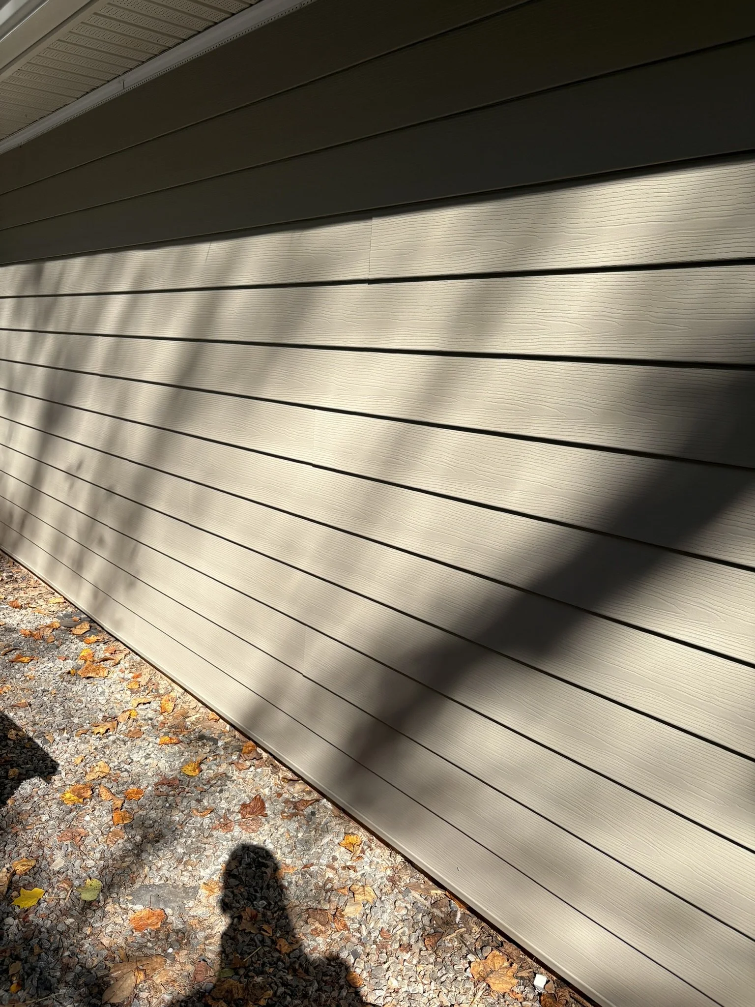 Close-up of beige horizontal siding on a building, with a small section of gravel and fallen autumn leaves on the ground at the bottom of the image, and shadows cast on the siding.