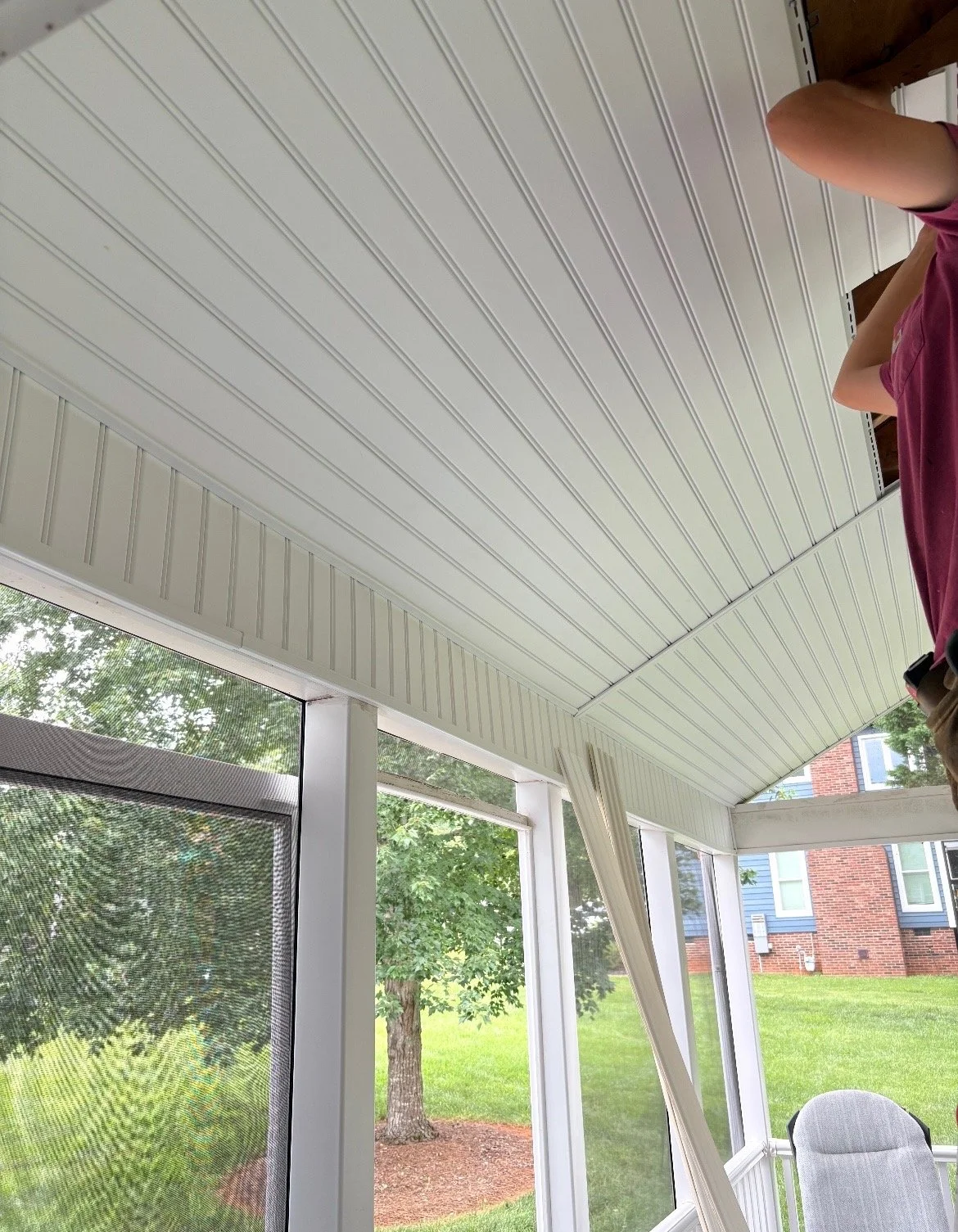 The image shows a screened-in porch with a white vinyl ceiling and window frames. The porch overlooks a yard with trees and grass, and a house with a brick exterior is visible in the background. A person is partially visible on the right side of the 