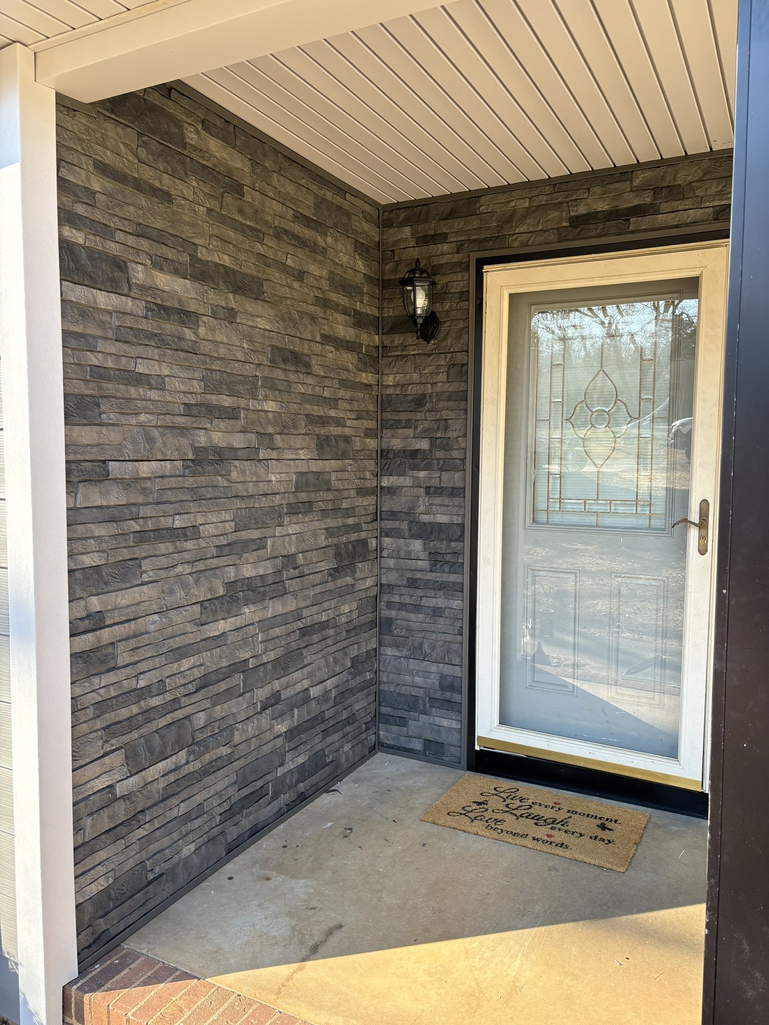 Front porch with dark stone wall, white trim, glass door with decorative design, black exterior light fixture, and a welcome mat.