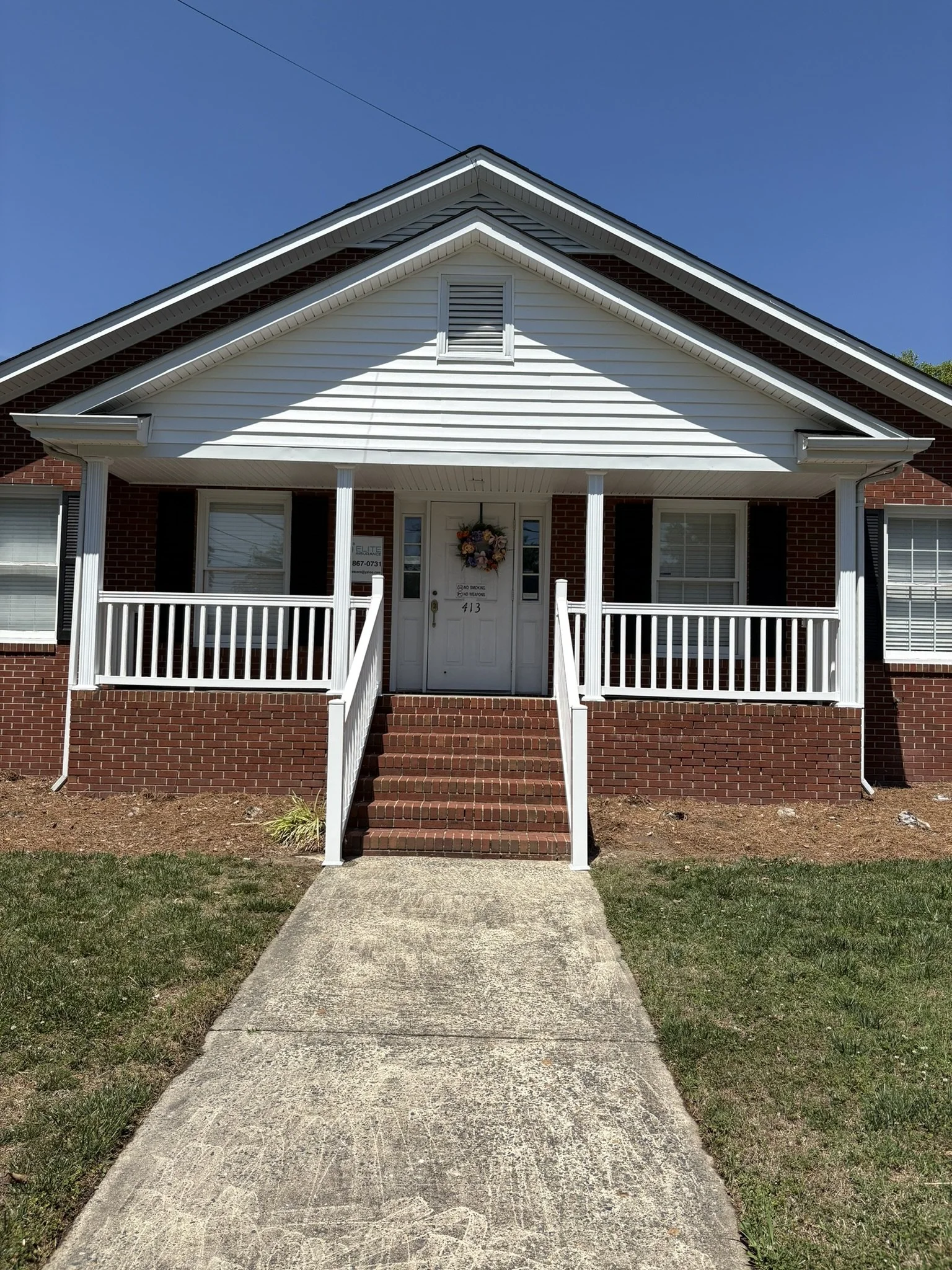 Front view of a house with a brick foundation, white siding, a small porch with white railings and steps, a wreath on the door, and a window on each side of the door, under a blue sky.