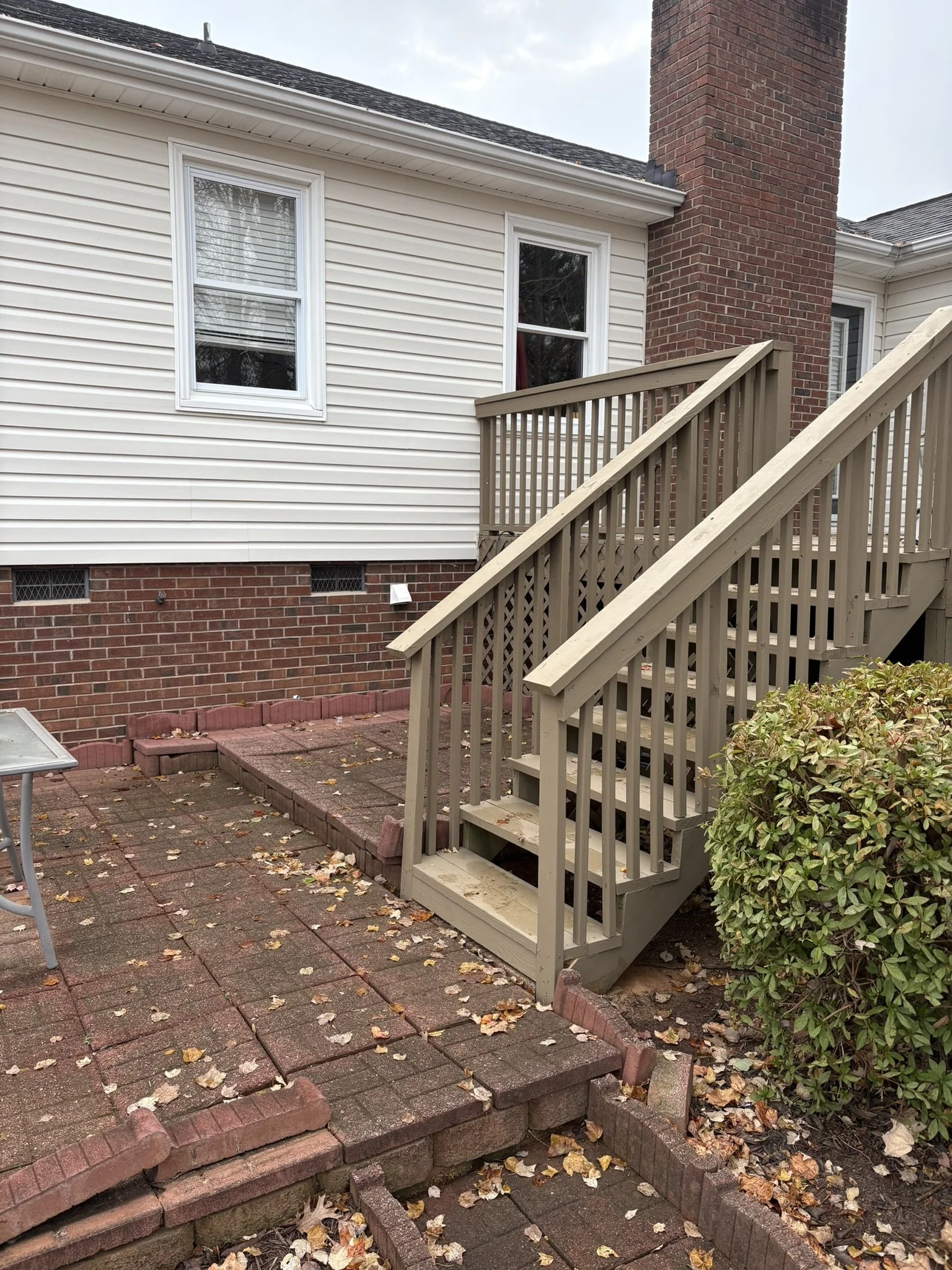 Backyard with a small set of wooden stairs leading up to a house door with white siding and a brick chimney, surrounded by some plants and fallen autumn leaves on the ground.