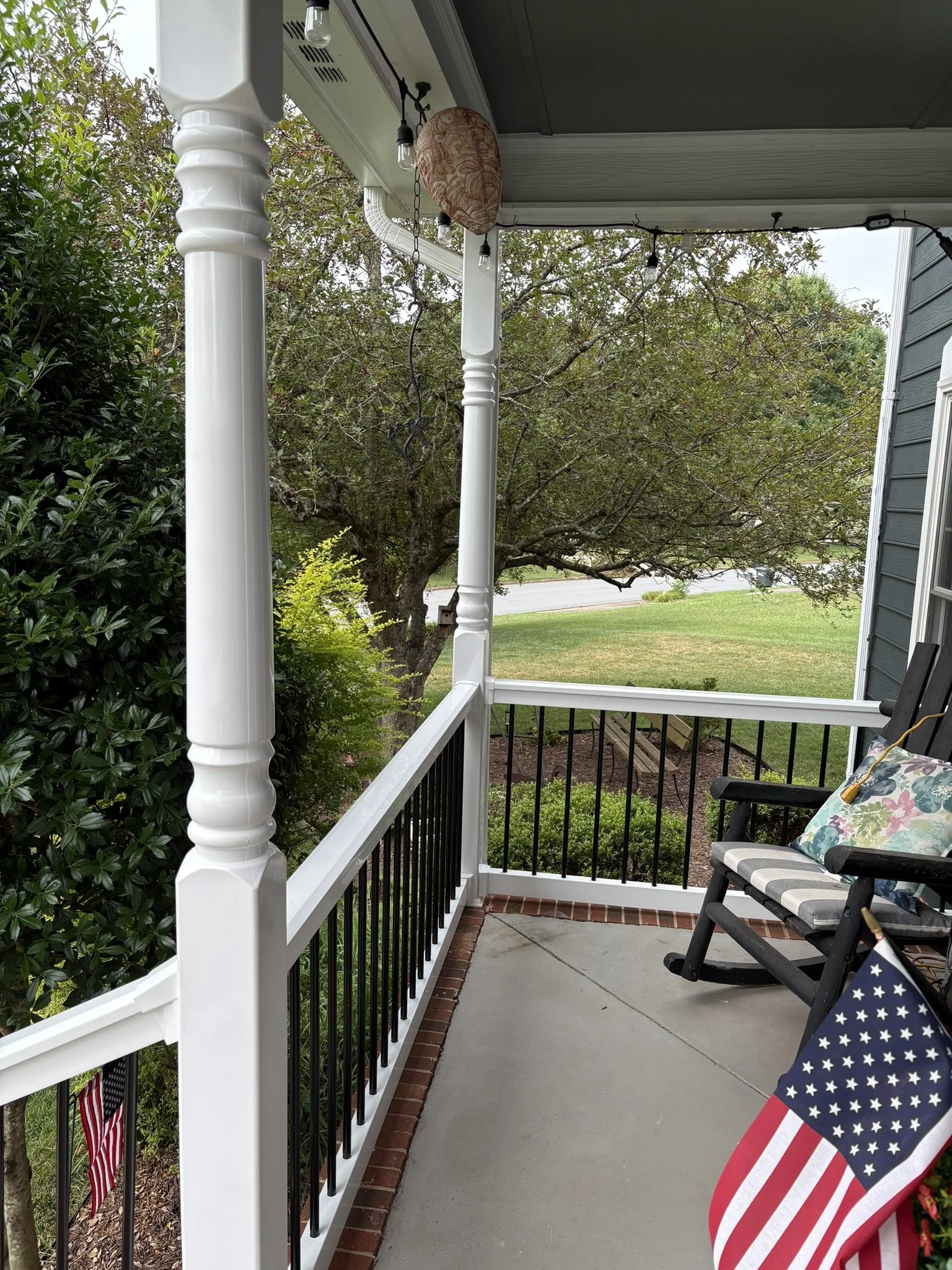 View of a residential porch with white railings, black balusters, and a chair with a patriotic star-spangled pillow, overlooking a yard with trees and lawn.
