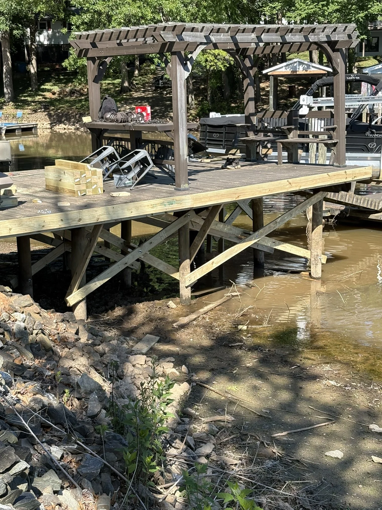 A wooden dock or pier extends above a small body of water, with construction materials and tools placed on it, including a stack of wooden planks, chairs, and a black backpack. There is a shade structure with a slanted roof on the dock. Trees and a h