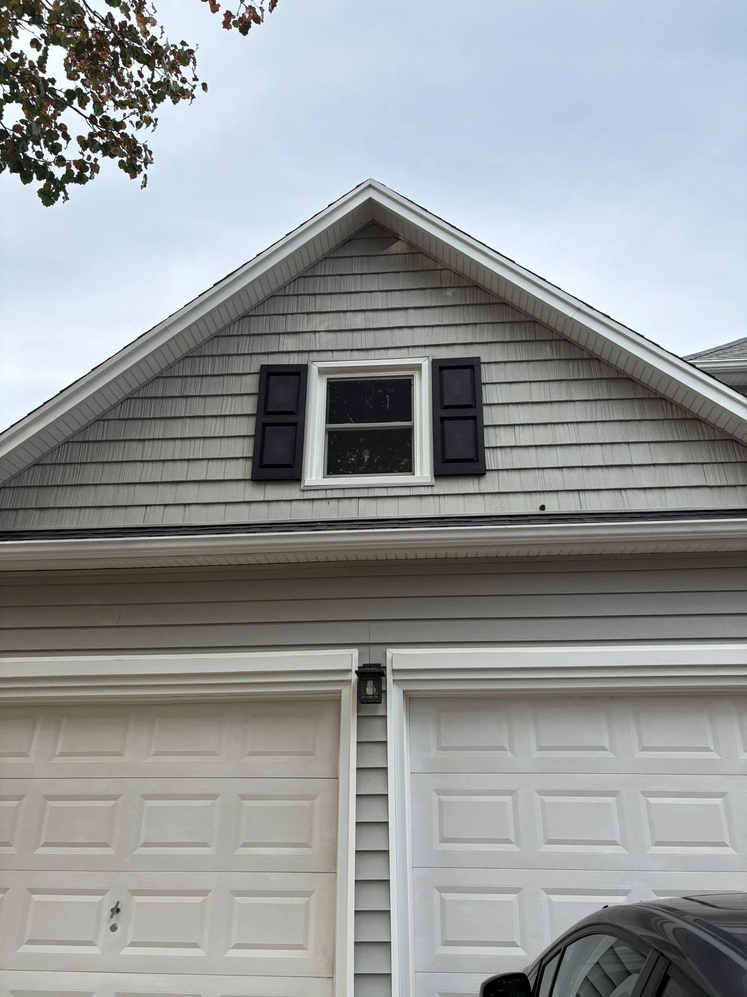 The image shows the upper part of a house with a gray gable roof and beige horizontal siding. There is a single window with black shutters on the gable wall. Part of a white garage door is visible at the bottom, along with a small section of a black 
