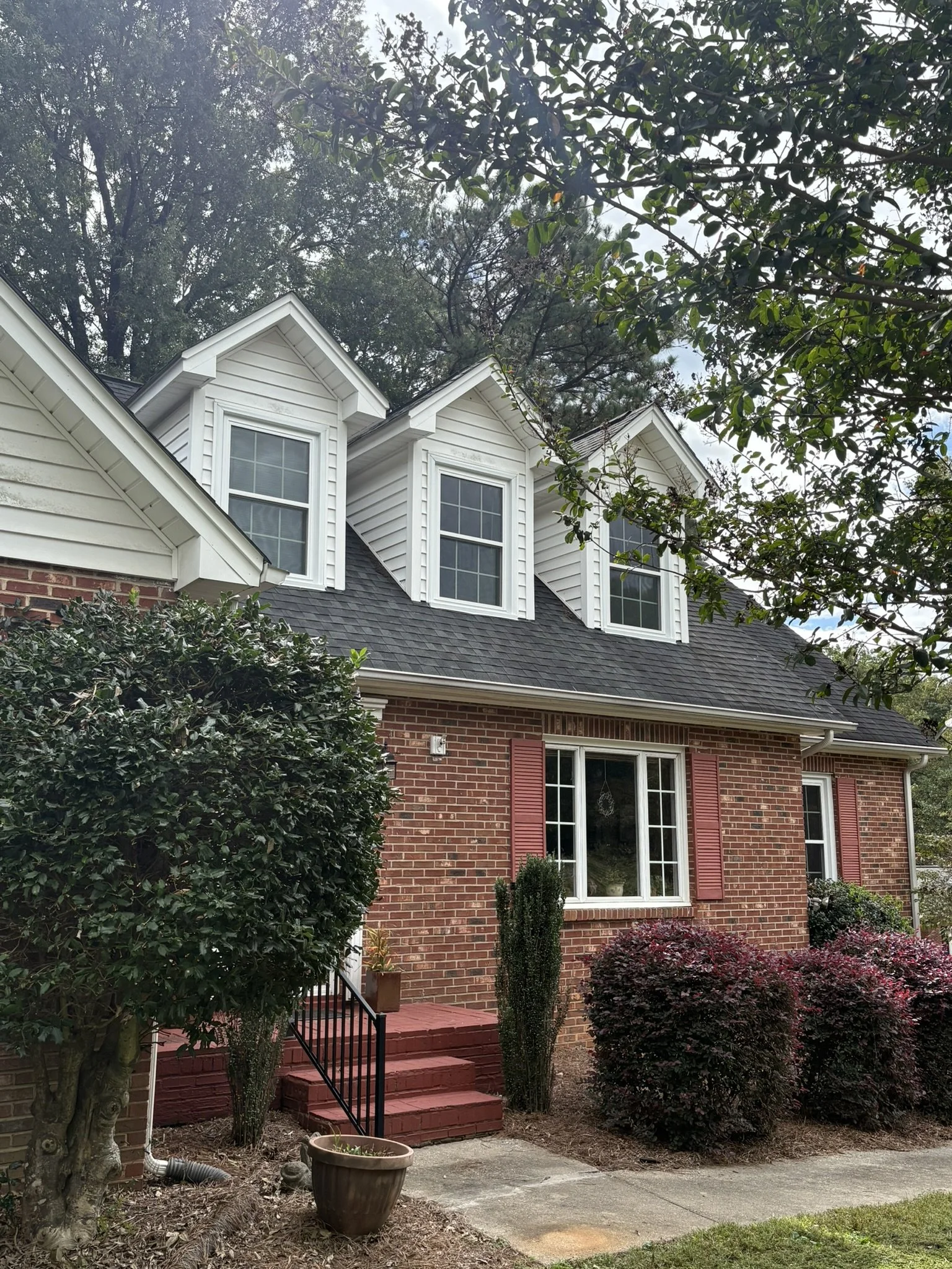 A two-story house with brick exterior walls, white window frames, and red shutters. The house has a steep dark shingled roof with three sawtooth dormer windows. There are several shrubs, small trees, and potted plants in the garden in front of the ho