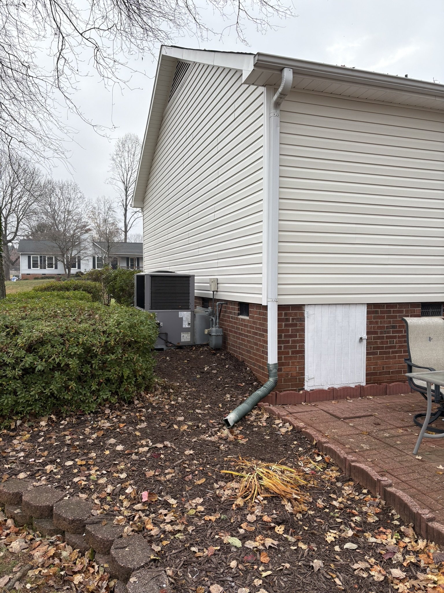 Backyard view of a house with white siding, a brick foundation, and an air conditioning unit. There is a gutter downspout, a garden area with fallen leaves, and part of a patio with red bricks. Some trees with bare branches are visible in the backgro