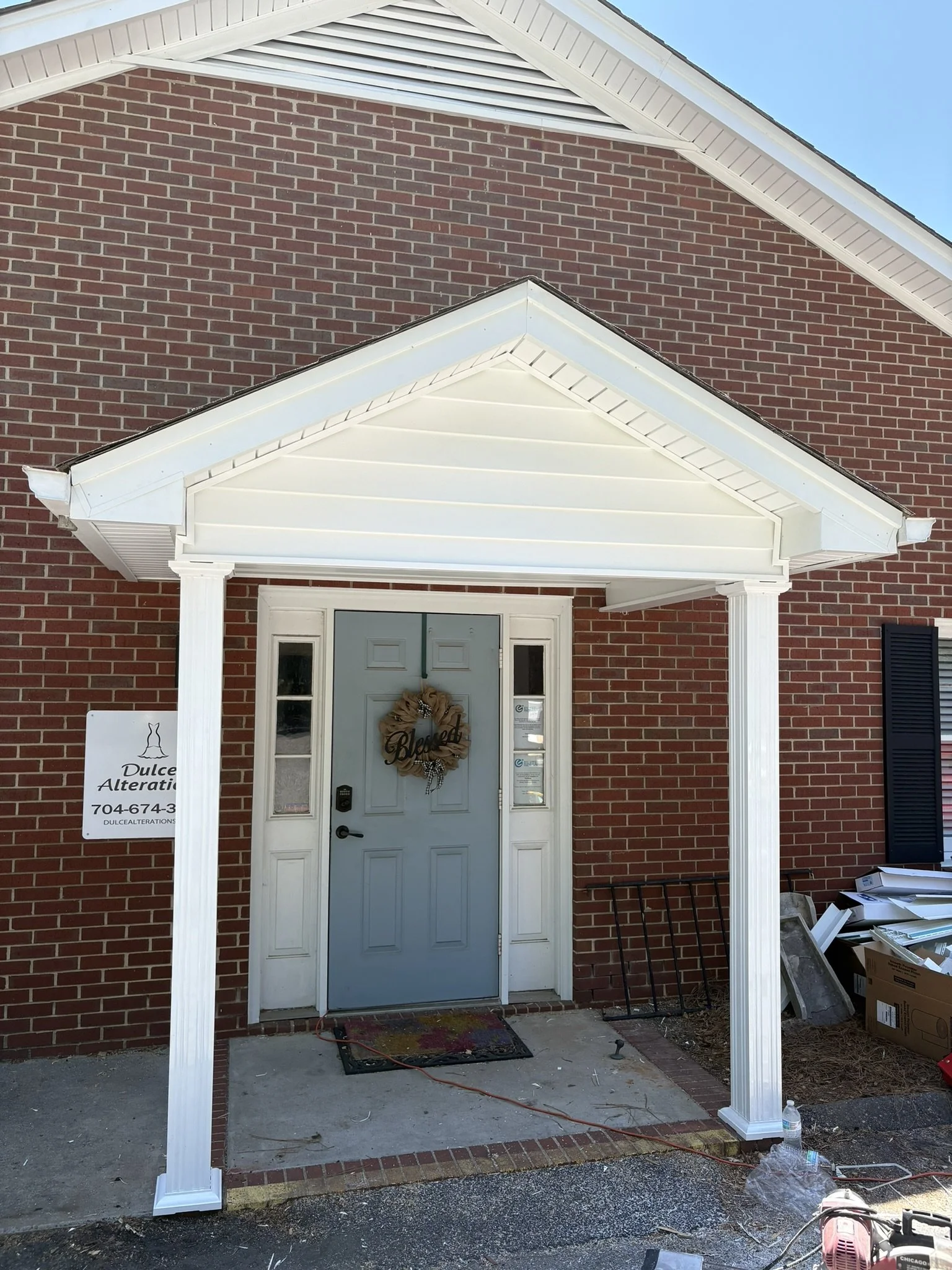 Front porch of a brick house with white pillars and a white decorative gable, with a front door decorated with a wreath, and construction signs and tools on the ground.
