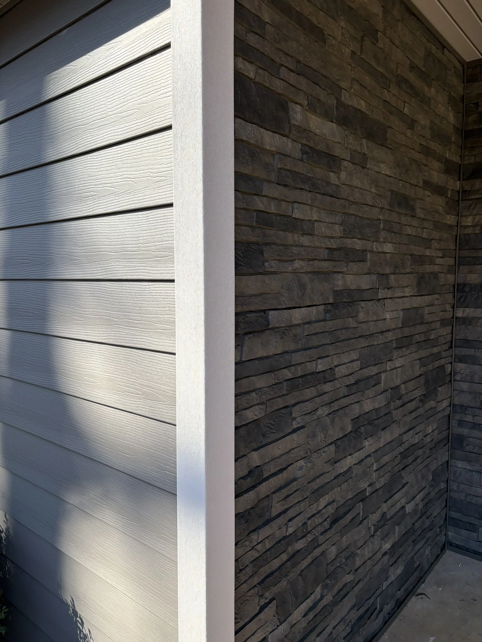 Exterior corner of a house showing transition from white horizontal vinyl siding to dark brown stone veneer.