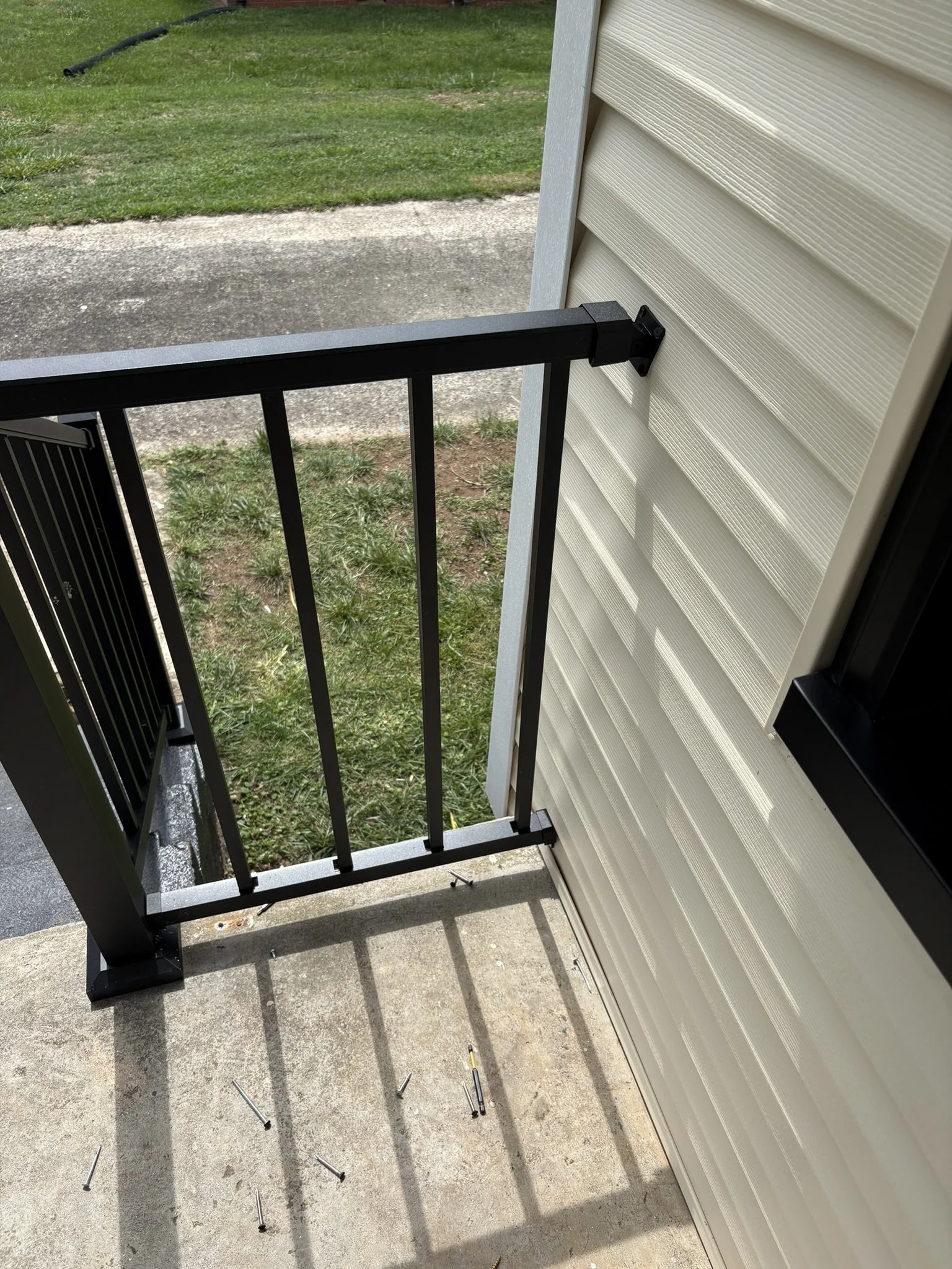 A black metal railing is being installed outside a beige vinyl-sided building. Several screws and tools lay on the concrete ground below the railing. The background shows grass and a paved area.