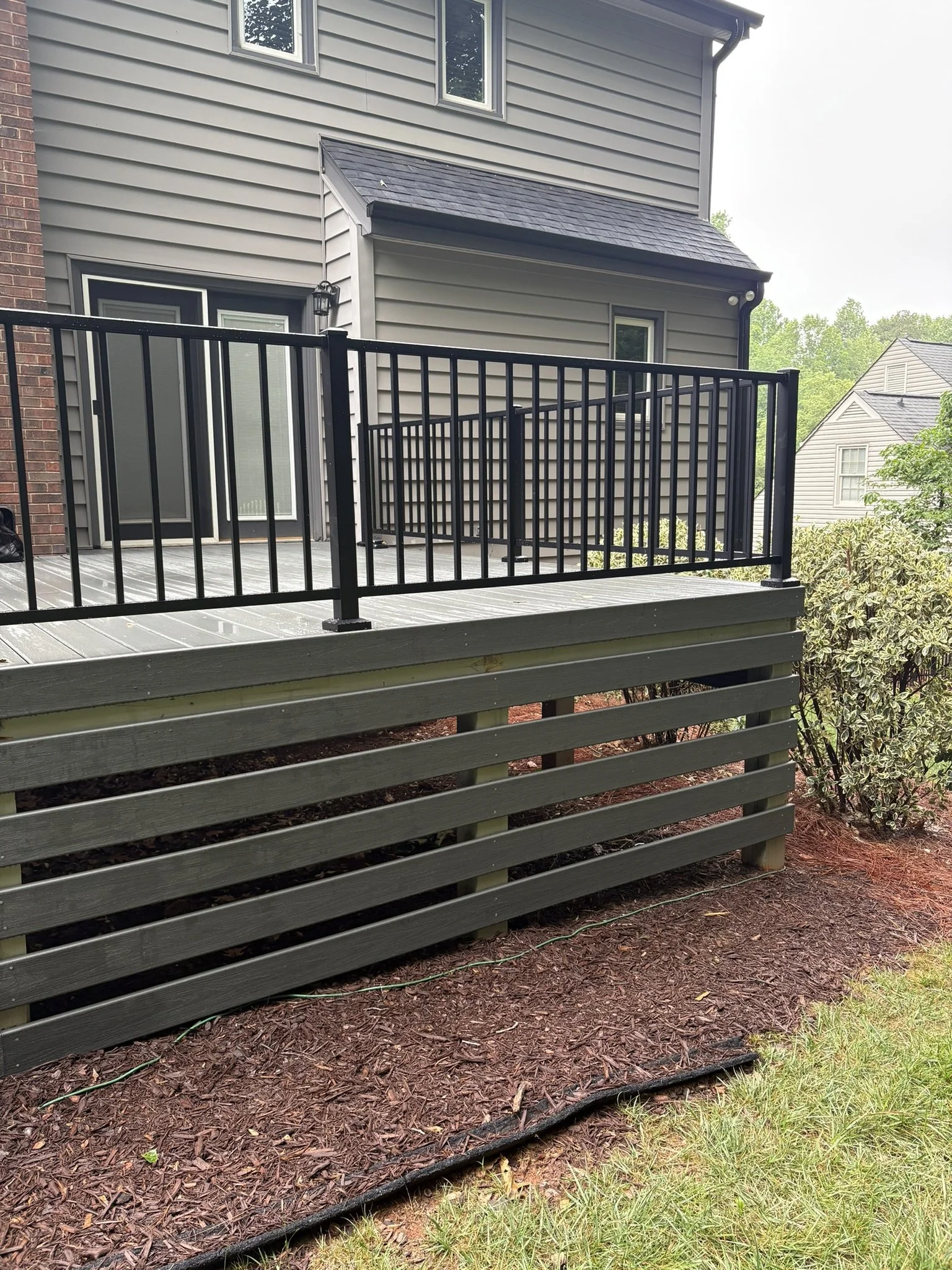 A backyard deck with black metal railing attached to a grey house with trim. There is a sliding glass door with blinds, a small window, and a porch light. The deck provides steps leading down to a mulched planting area with some bushes and grass. The