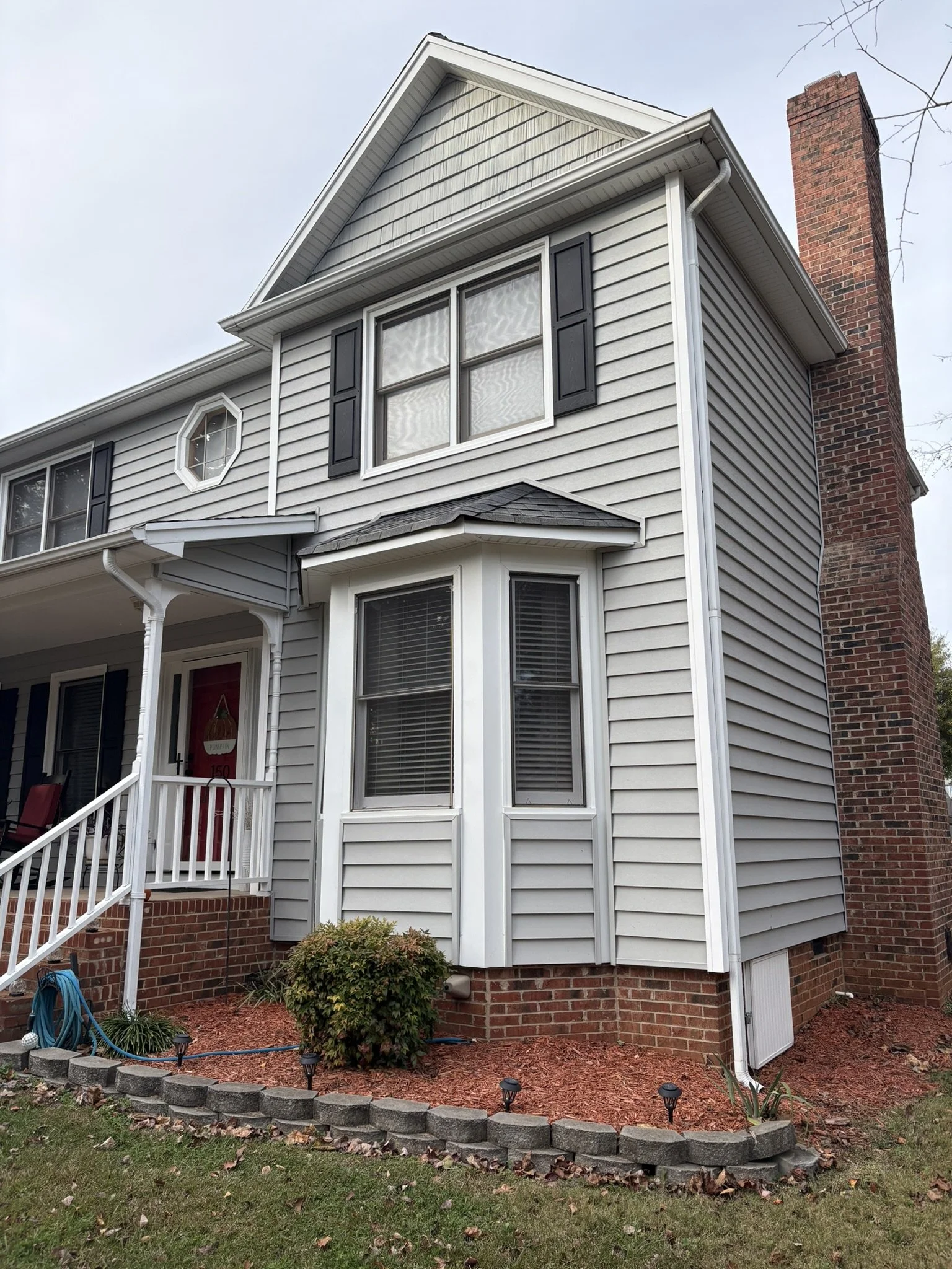 The front view of a two-story house with gray siding, white trim, and black shutters. The house has a brick chimney on the right side, a bay window on the lower level, and double-hung windows on the upper level. The front porch has a white railing an