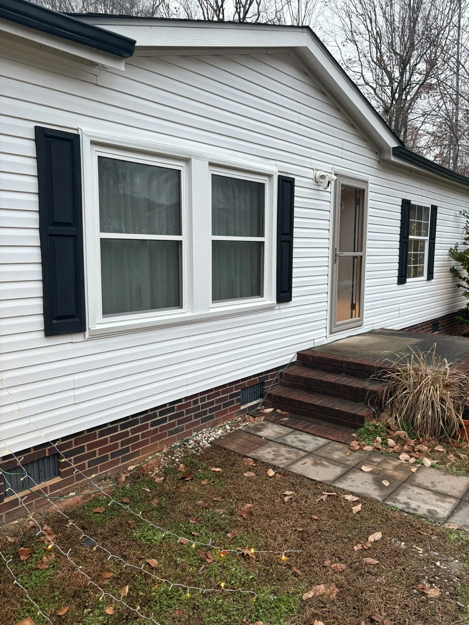 A house with white vinyl siding, two double-hung windows with black shutters, a glass storm door, brick stairs leading to the door, and a small yard with patches of grass, fallen leaves, and a string of outdoor lights.