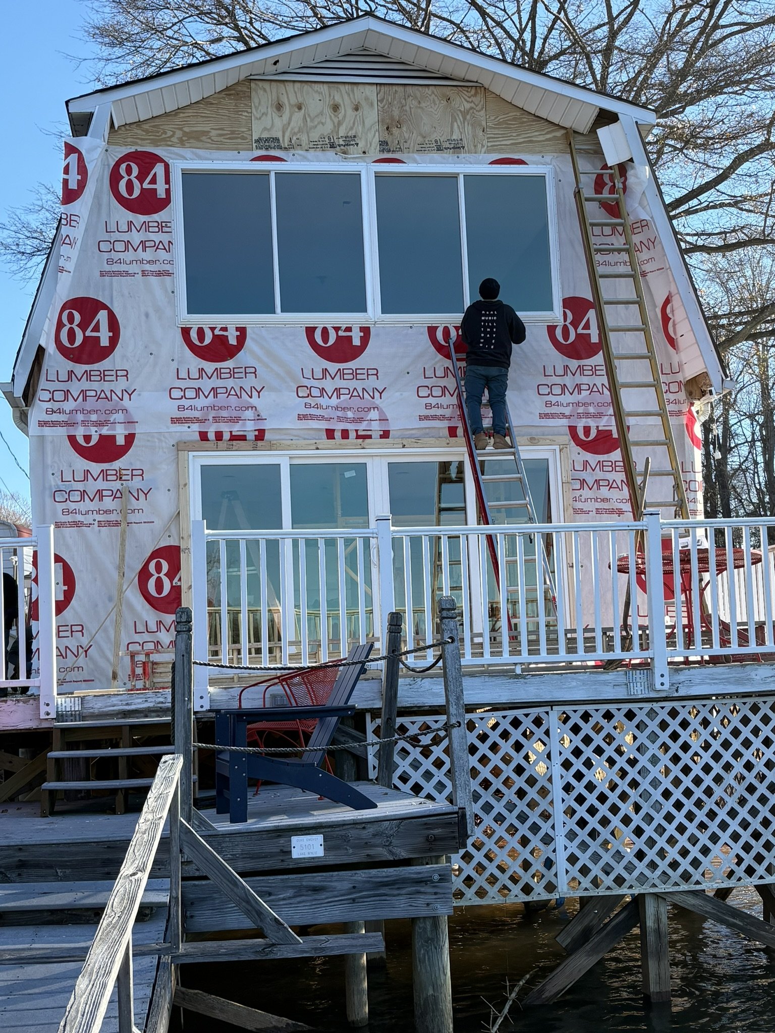 A house under construction with the exterior wrapped in insulation material, and a large window installed. A tall ladder is leaning against the house, and a person is standing on a small extension ladder, working on the window. The house is elevated on stilts over water, with a wooden staircase leading up to a porch. There are trees in the background and a clear blue sky.