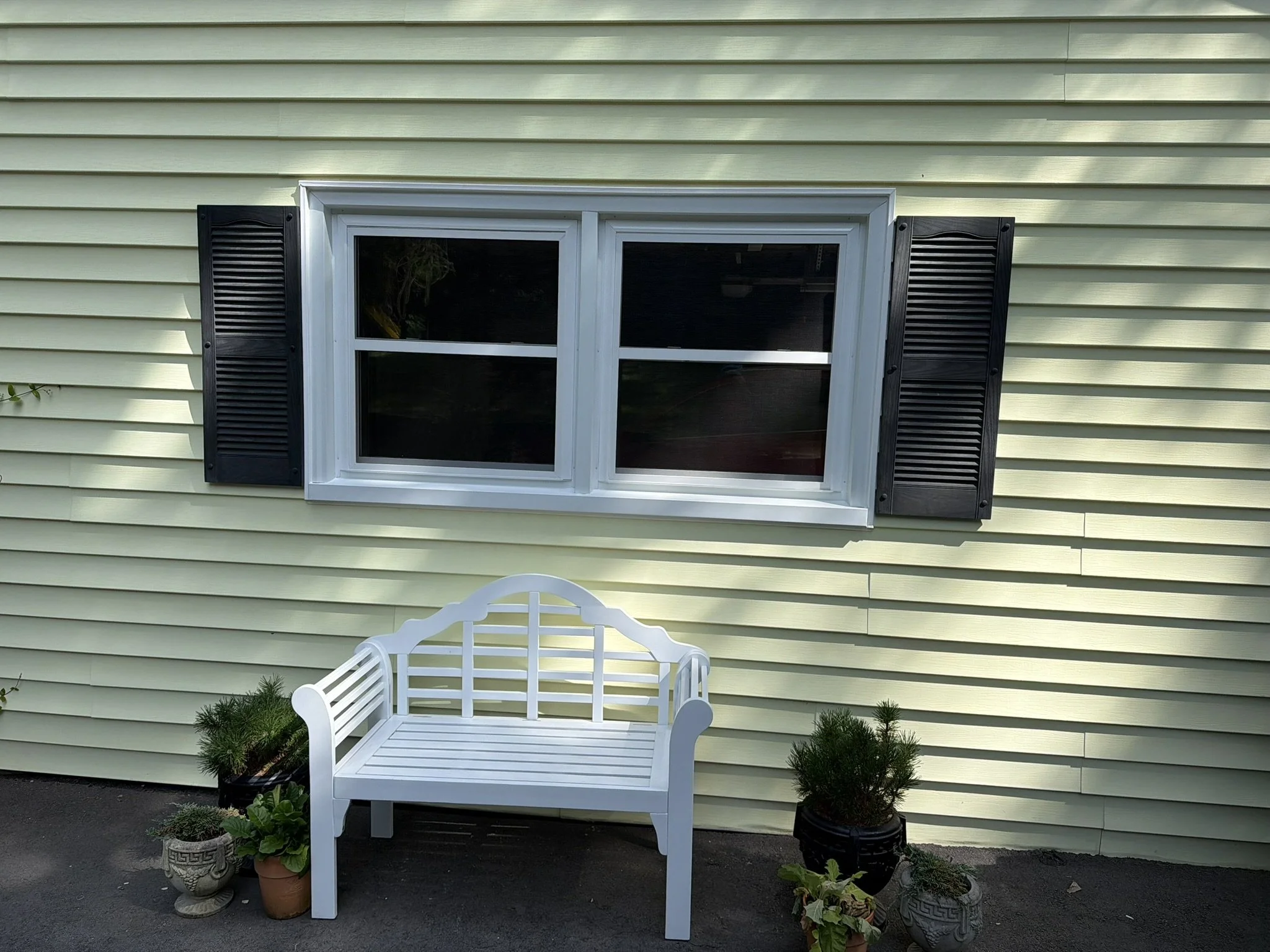 White outdoor garden bench in front of a house with light yellow siding, surrounded by potted plants, near a window with black shutters.