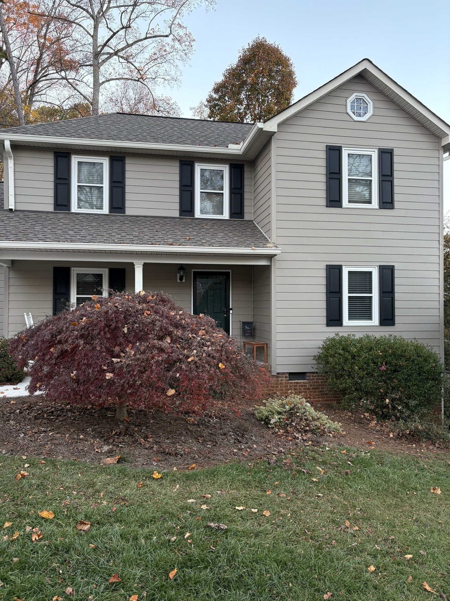 Front view of a two-story house with beige siding, black window shutters, a front porch with a pillar, and various shrubs and a tree in the yard with fallen leaves.