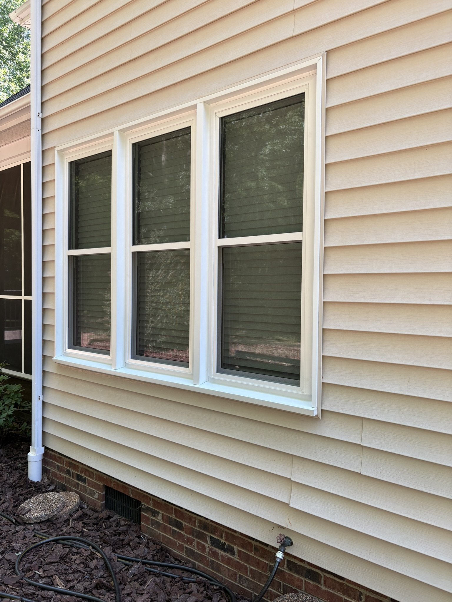 Residential house with beige vinyl siding and three double-hung windows with white frames, partially reflected in the glass, with a brick foundation and garden hoses on the ground.