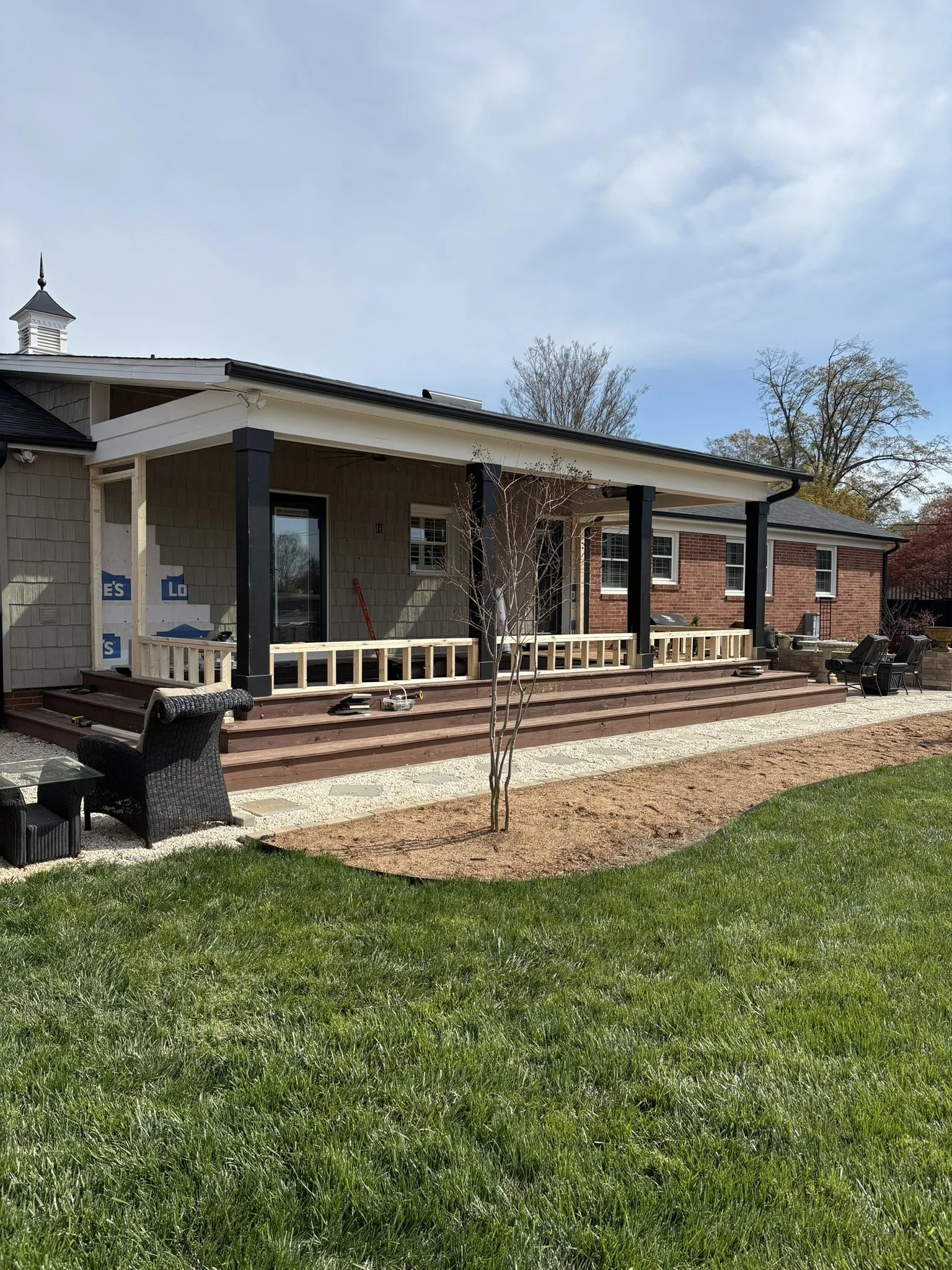 A house undergoing construction or renovation with a new deck being built. The house has beige siding, black columns, and a brick section. There are construction tools and materials on the deck, and a small tree in the yard. The sky is partly cloudy 