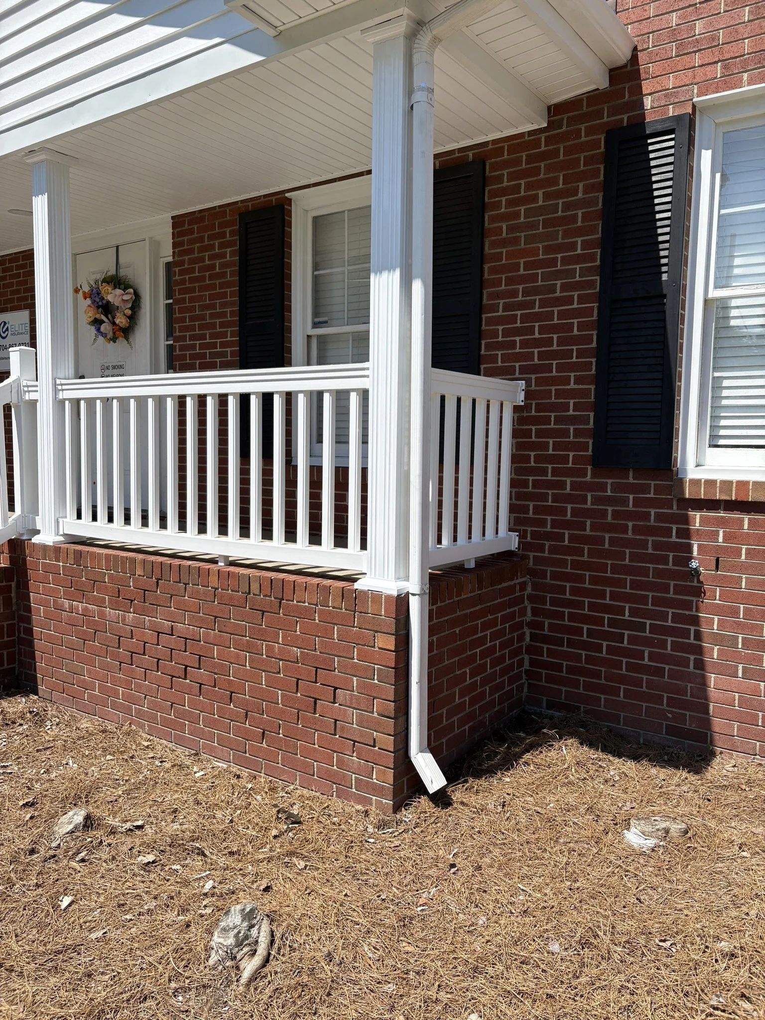 Close-up of a brick porch with a white railing and support columns, a window with white trim, black shutters, a decorative wreath on the door, and an outdoor water faucet with a hose connection, on a sunny day with dry grass and pine needles on the g
