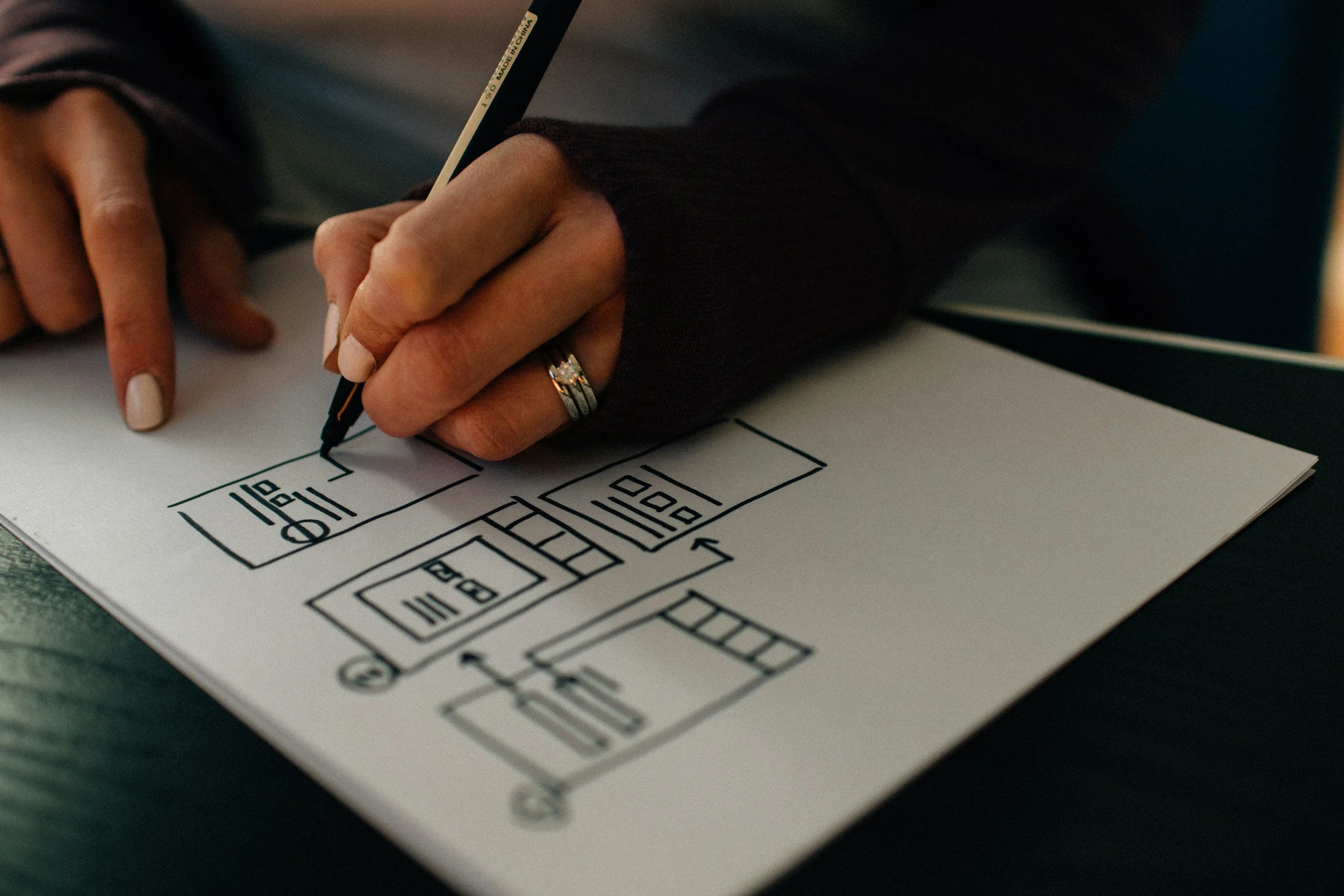 A person wearing a black long sleeve shirt is drawing a floor plan or blueprint on a piece of paper with a black marker. The paper shows various room layouts and labels.