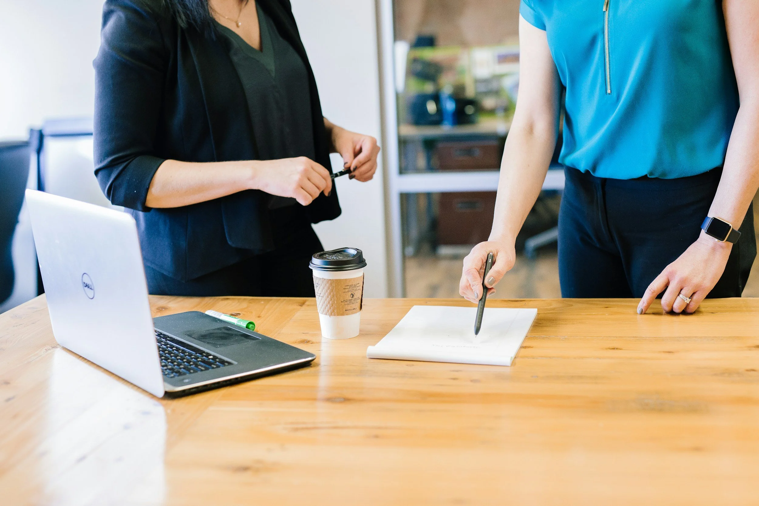 Two women are working together at a wooden table with a laptop, a takeaway coffee cup, a green marker, a notepad, and a pen. One woman is wearing a black blazer and the other is in a blue top and dark pants. They are in an office or meeting room with shelves in the background.