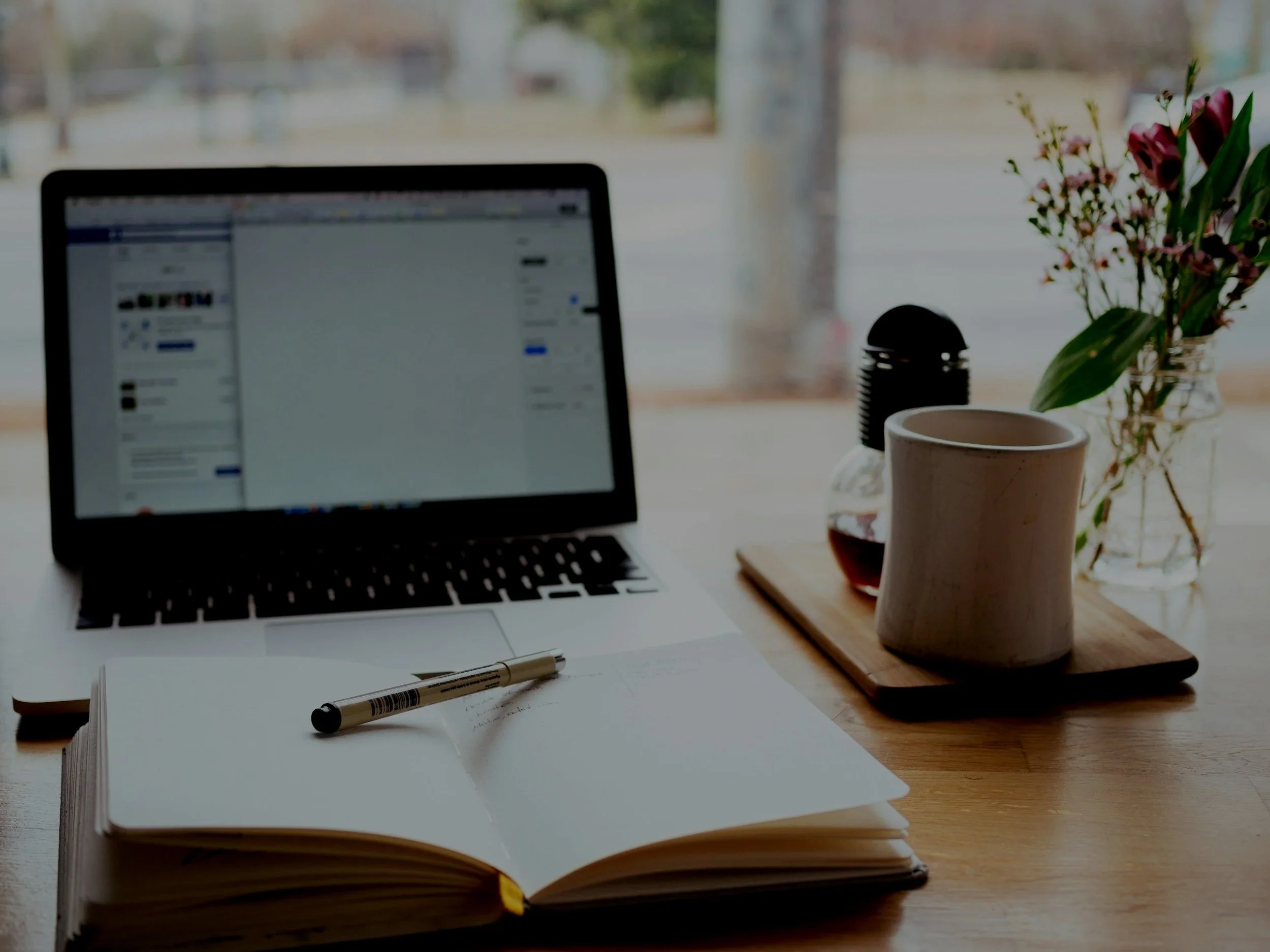 Open notebook with handwritten notes and a pen on a wooden desk. In the background, a laptop displaying a social media page, a coffee mug, a glass of water, a pen, and a small bouquet of flowers in a vase near a window with trees visible outside.