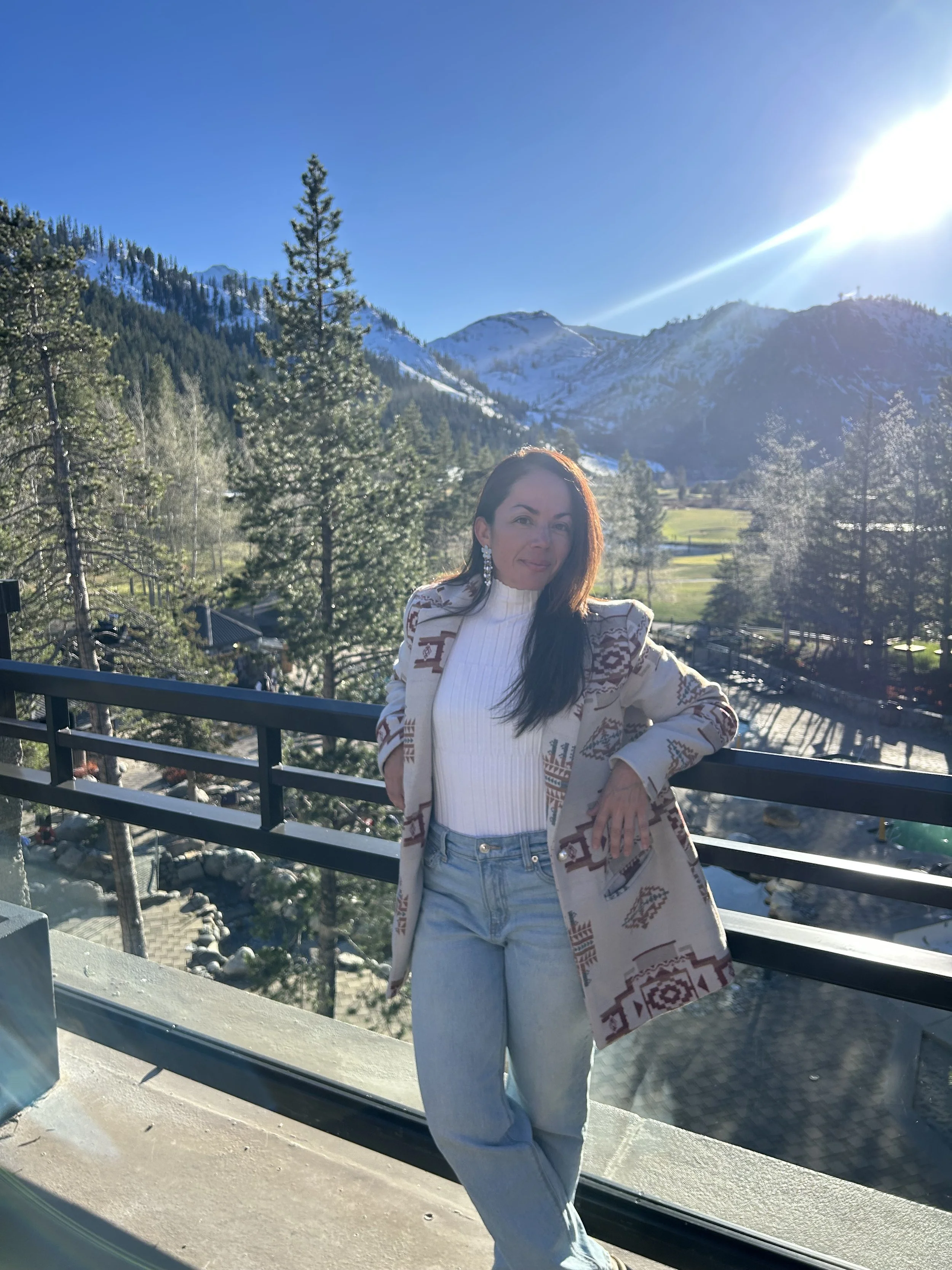 A woman with long dark hair, wearing a white turtleneck, light blue jeans, and a beige patterned coat, standing on a balcony with mountains and trees in the background on a sunny day.