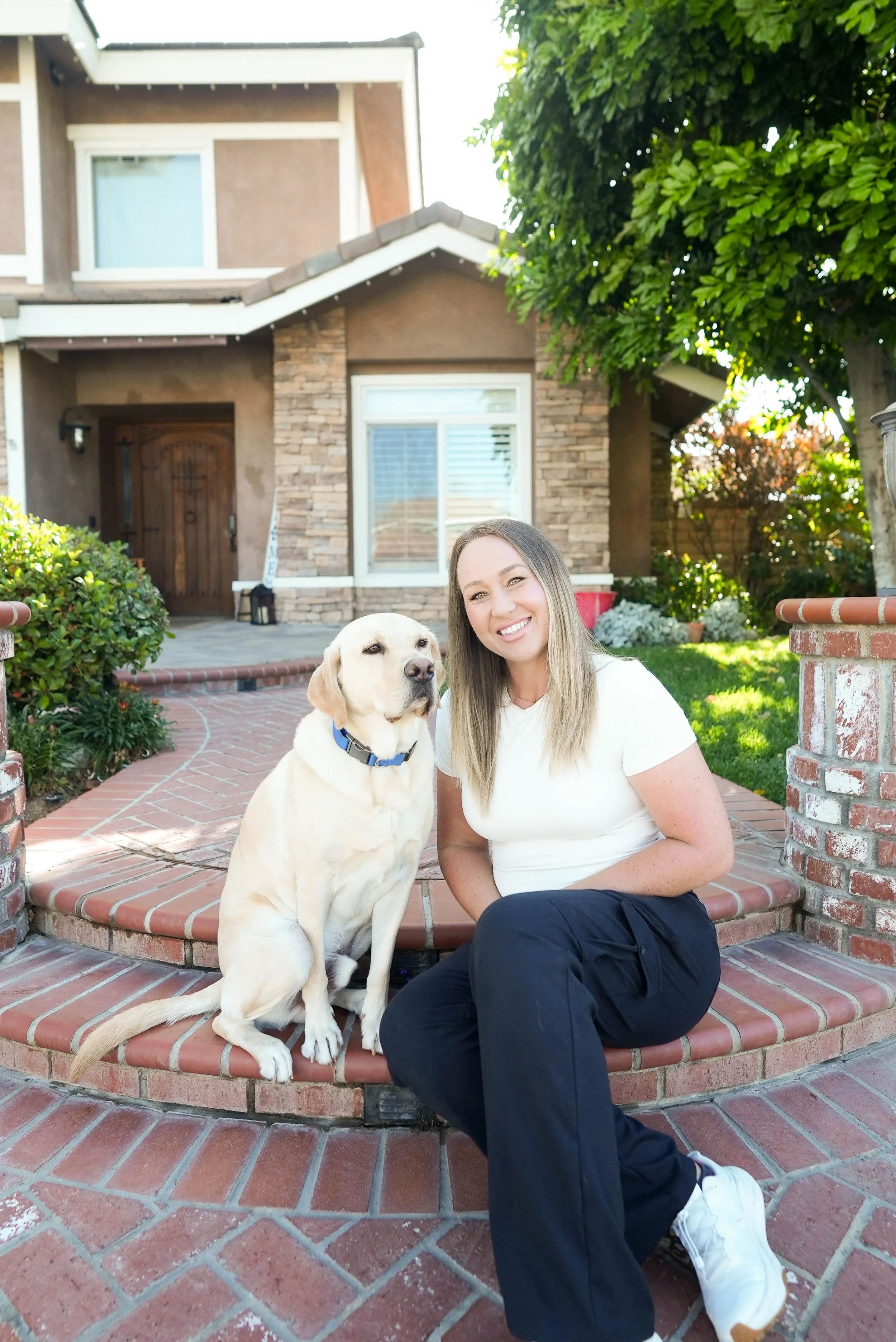 A woman smiling and sitting on brick stairs outside a house with a Labrador retriever dog. The house has a front door, a window, and is surrounded by greenery.