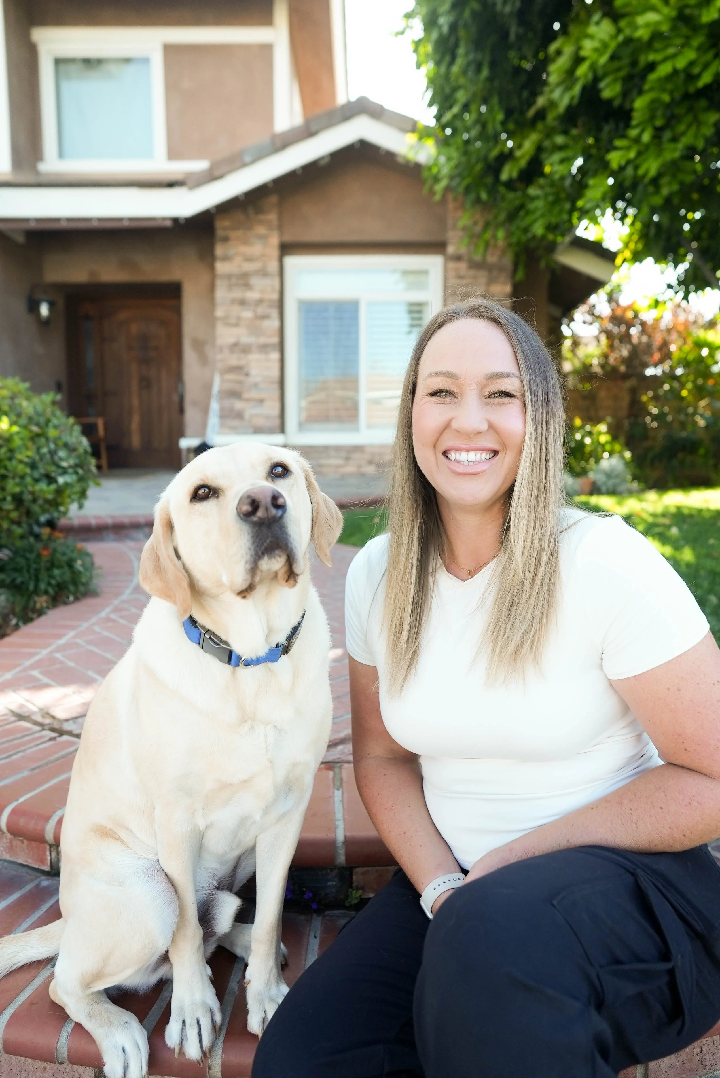 A woman with long blonde hair smiling, sitting on steps outside a house with a yellow Labrador Retriever dog beside her. The house has a brick and brown exterior with a large window and green trees around.