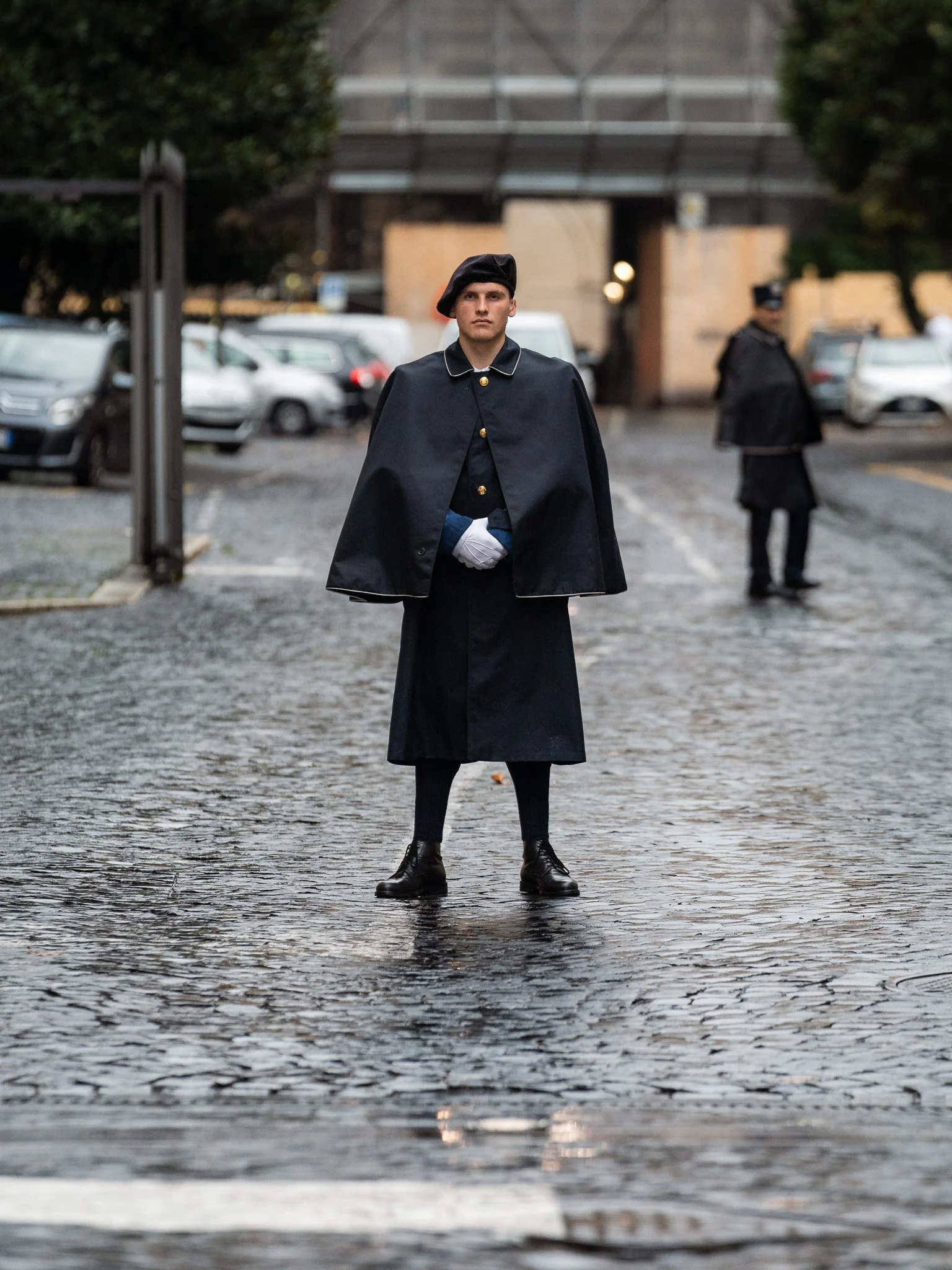 The Gendarmerie officer standing alone on wet cobblestones in the rain, cape, white gloves, another figure echoing behind him. Cinematic and still. Feels like a film frame.