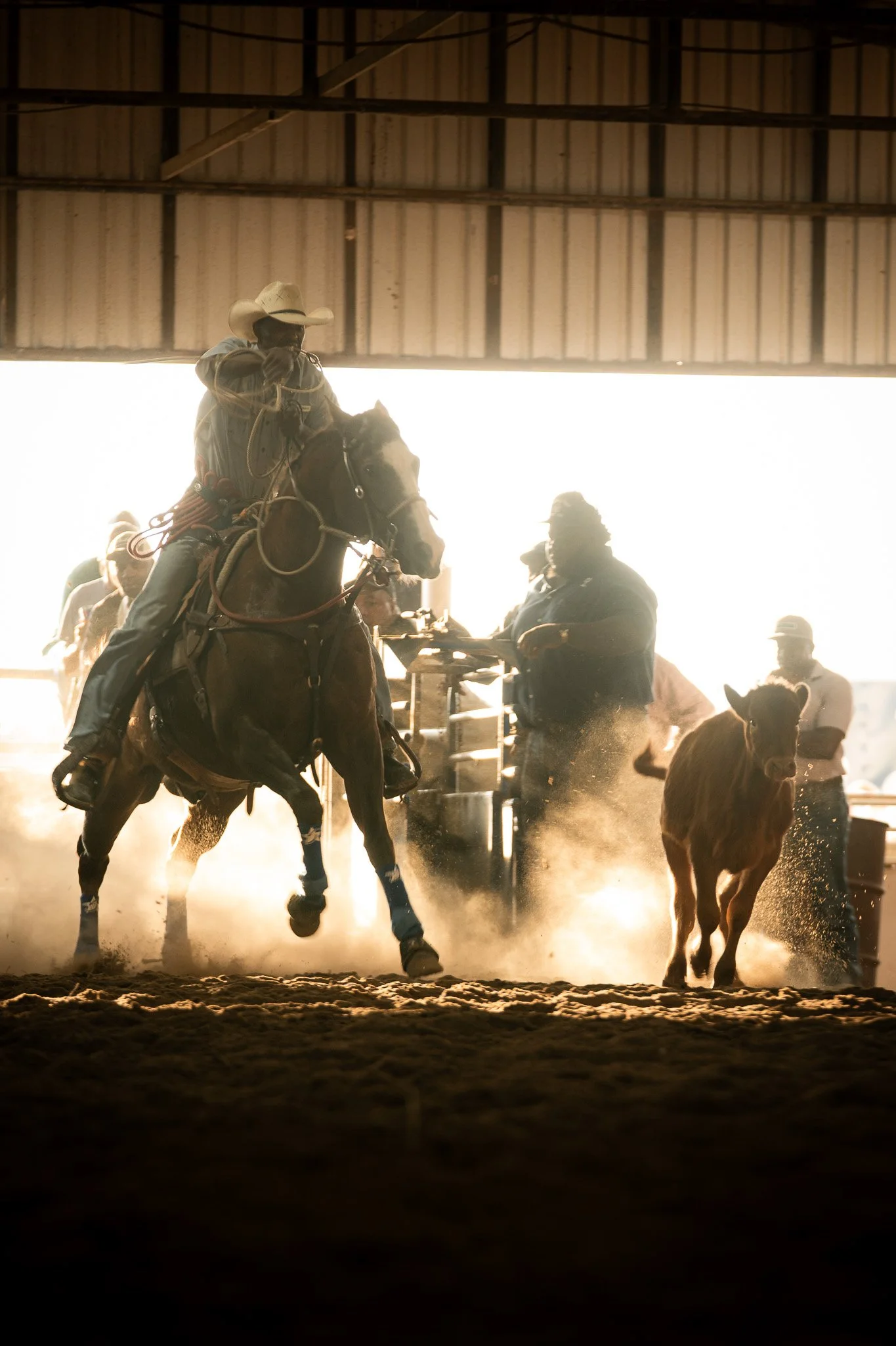 Riders on horses and people on foot in a dusty indoor rodeo or horse riding arena, with a metal roof overhead and backlighting from the bright outside shot in November of 2024 for Jas Prince's Rodeo Birthday in Hempstead, Texas.