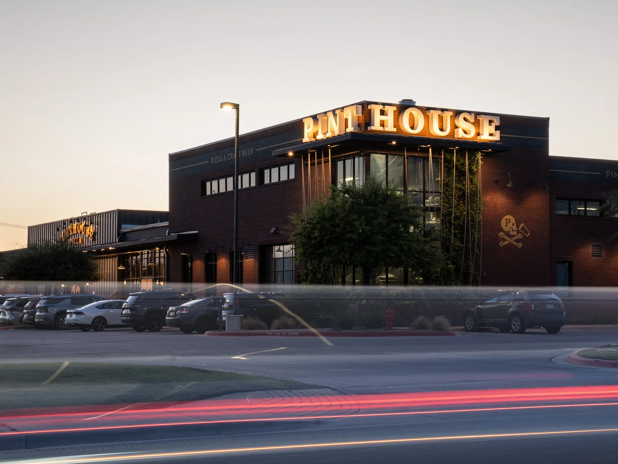 Exterior of a brick building with a large illuminated sign that reads 'PINT HOUSE' against a clear evening sky. There are parking lot cars in the foreground and light trails from moving vehicles.