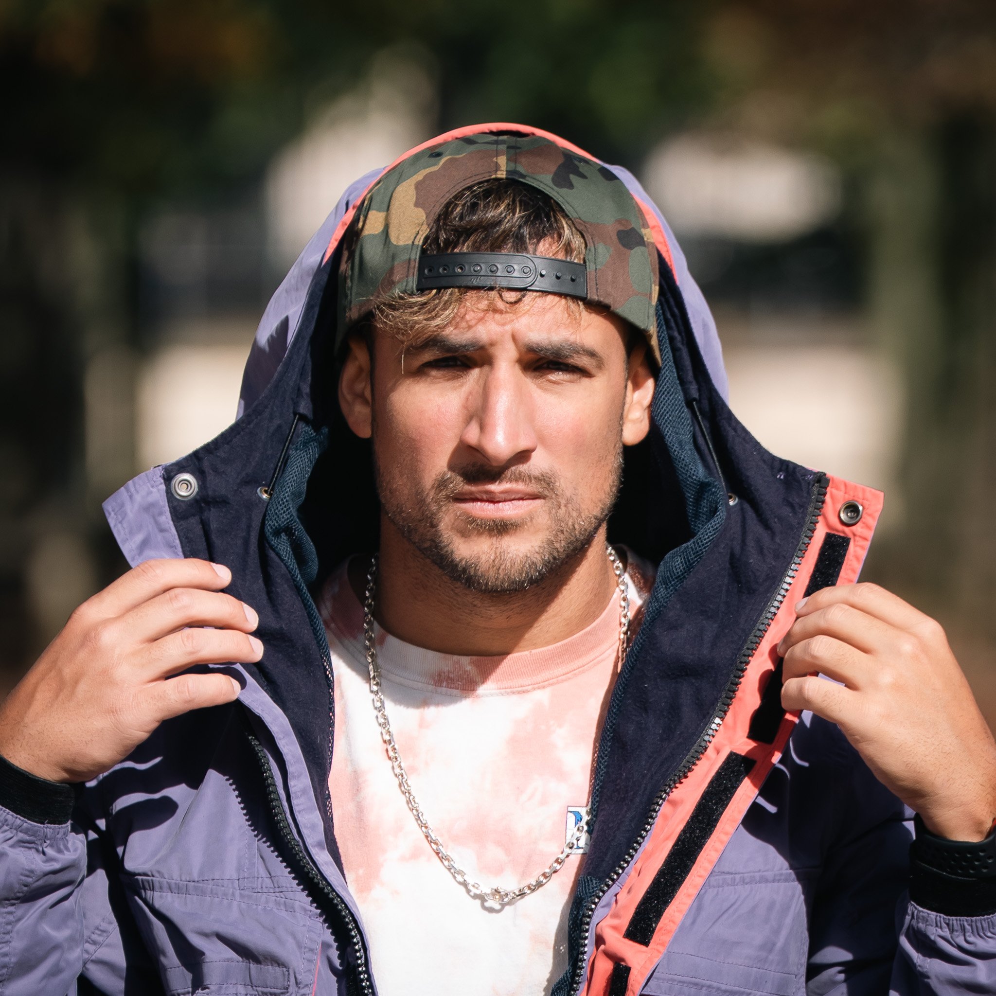 Close-up of Carlos in Paris, France wearing a camouflage baseball cap backward, a purple Abercrombie & Fitch jacket, a pink shirt, and a silver Tiffany and Co. necklace, outdoors with blurred background.