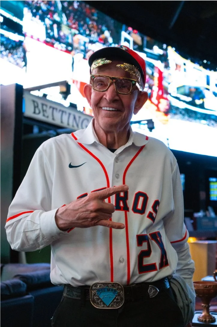 An elderly man wearing a Houston baseball jersey, a cap, and glasses, standing in a sports bar. He is smiling and making a rock and roll hand gesture, with a betting stand and TV screens behind him.