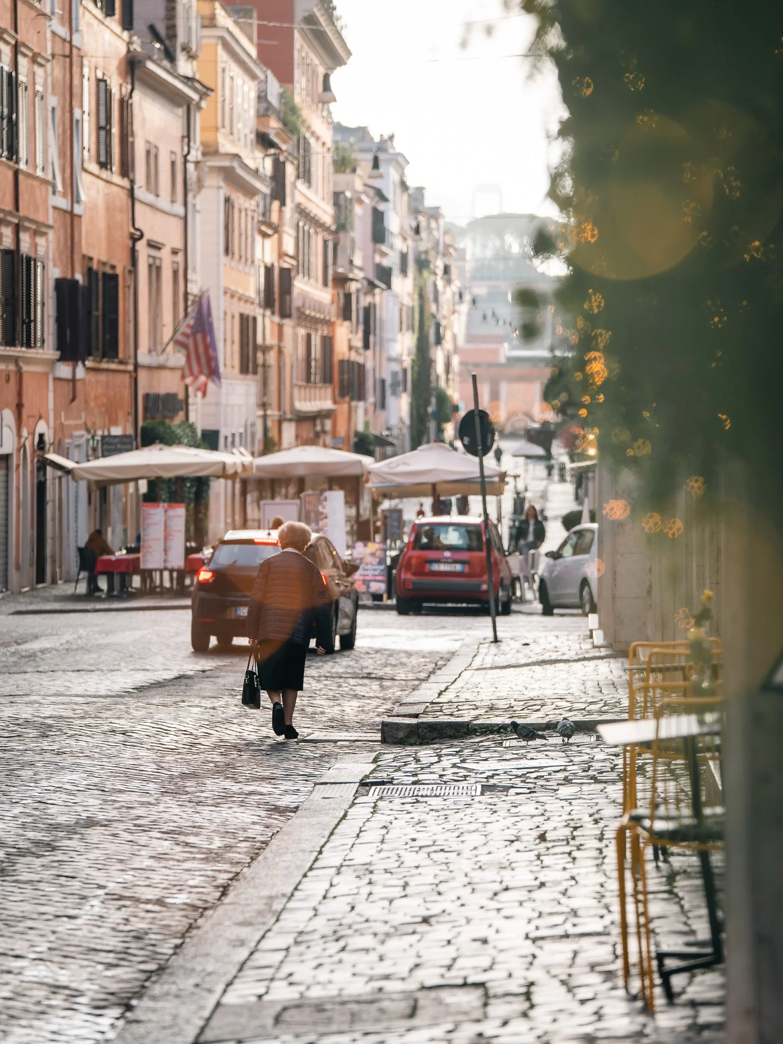 Street view in Rome, Italy with cobblestone pavement, historic buildings with colorful facades, outdoor cafe tables with umbrellas, and a woman walking away from the camera. The shot captured in the early morning hours.