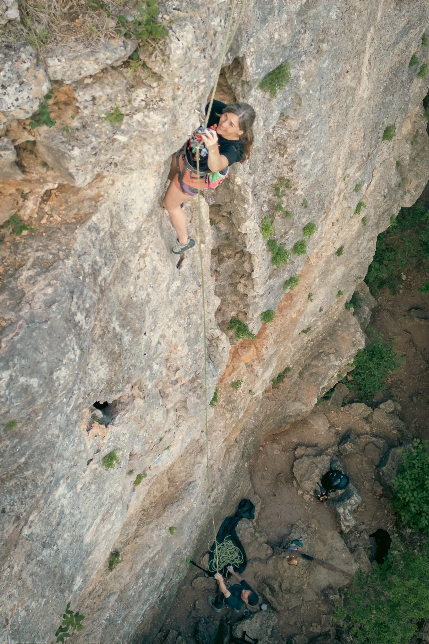 A woman rock climbing on a vertical rocky surface with climbing gear and harness, taken from above with a DJI Mavic 3 Drone. Shot at Reimers Ranch in Texas.