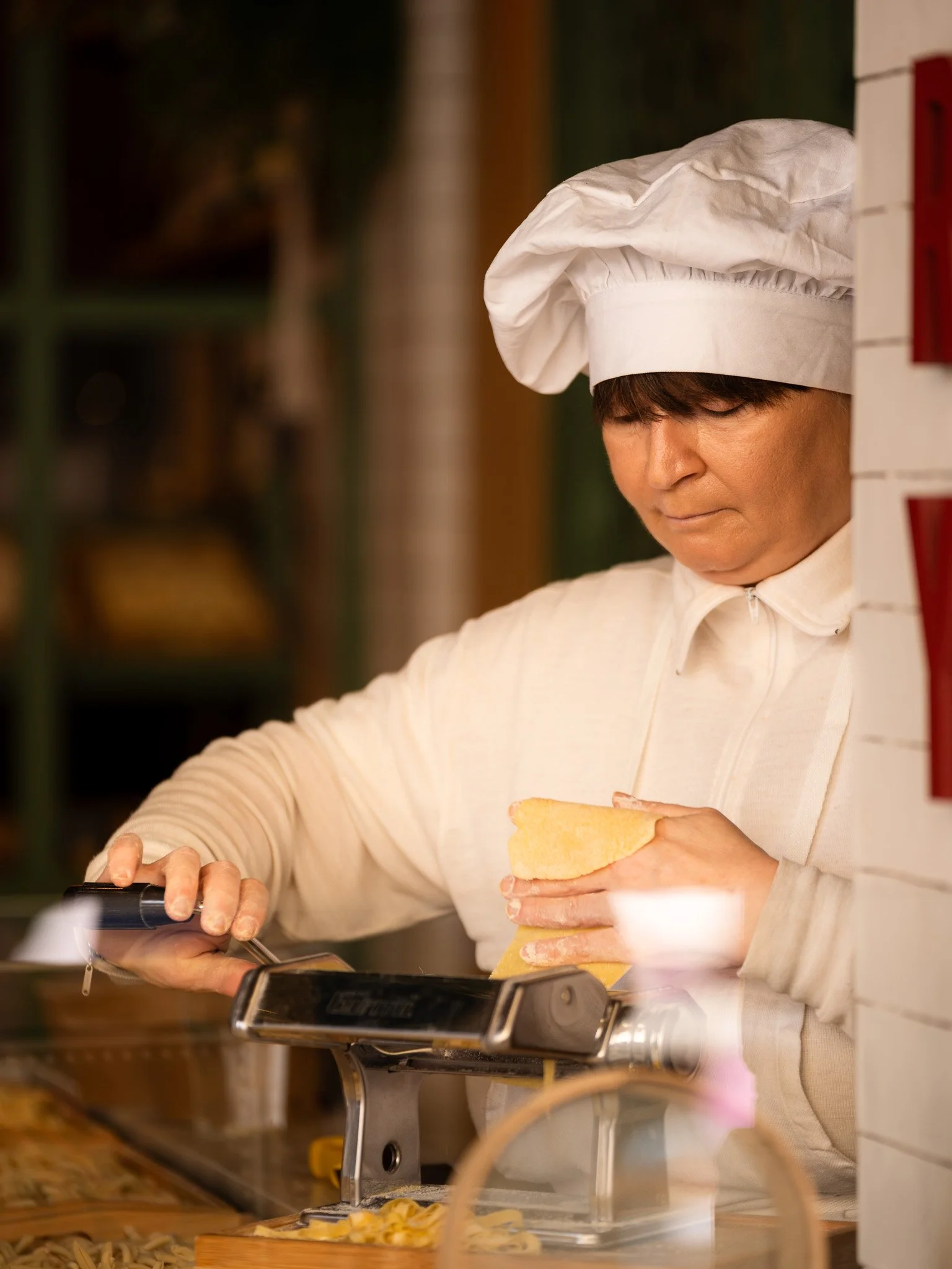An Italian woman pasta maker wearing a white chef hat and white jacket, photographed through a window, holding pasta in one hand and using tongs with the other.