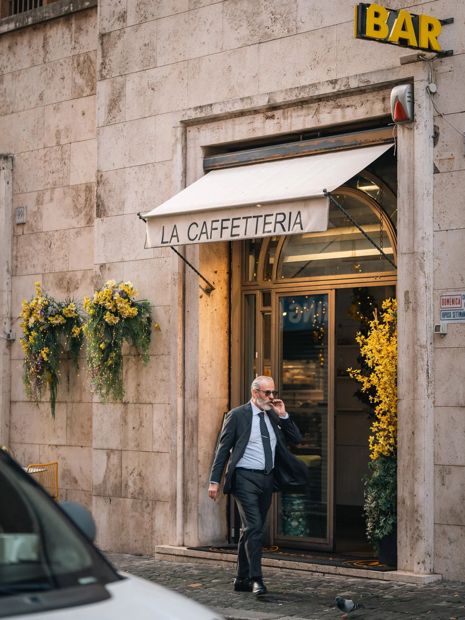 A man in a suit and sunglasses walking on a cobblestone street in front of a cafe with a white awning that says "LA CAFFETTERIA". There are yellow flowers in pots on either side of the entrance. A pigeon is on the ground near him, and part of a car i