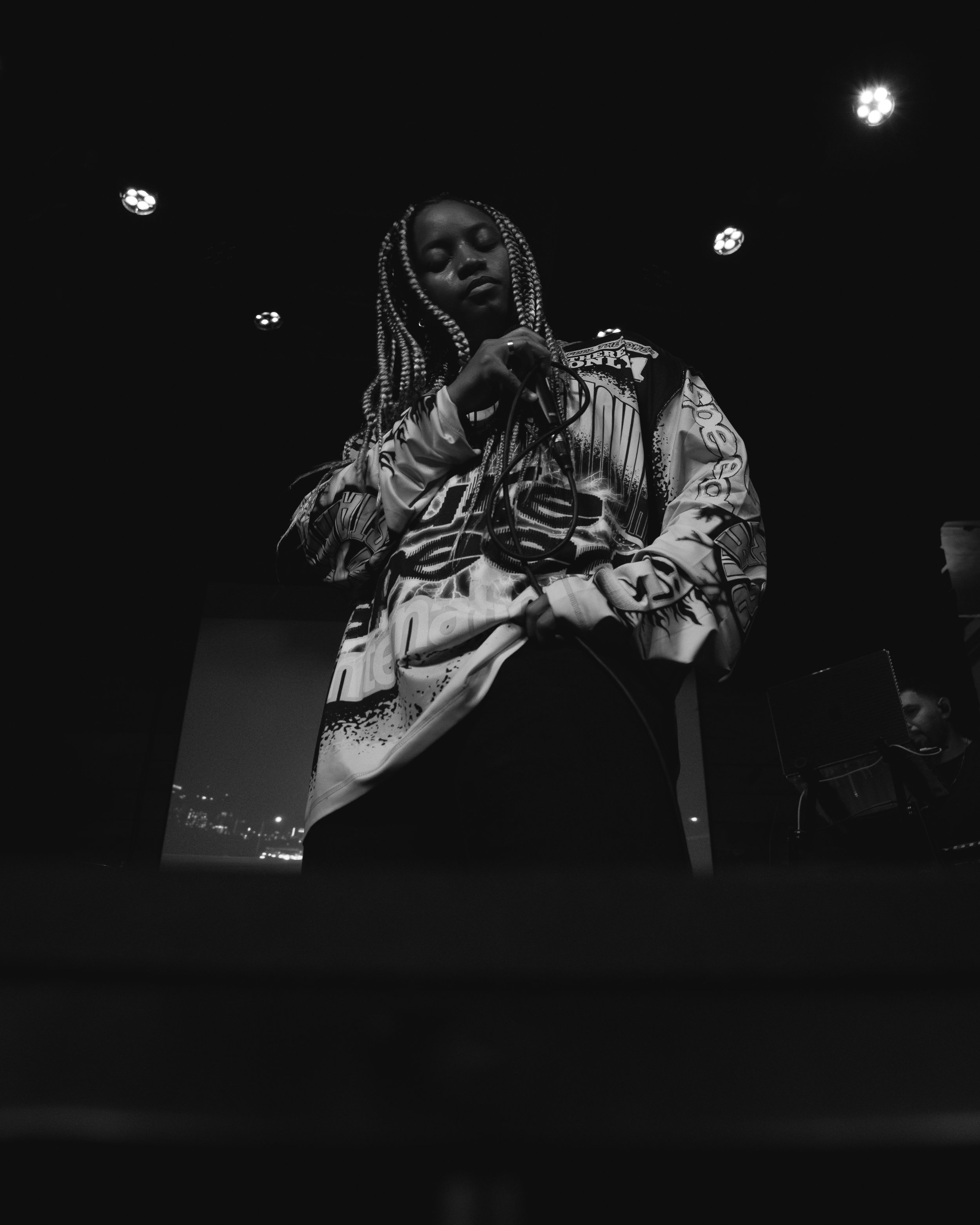 Artist from Magna Carda, photographed during a still moment during the concert at Mohawk Austin, with braided hair singing or speaking into a microphone on stage in a dark setting, with stage lights overhead.