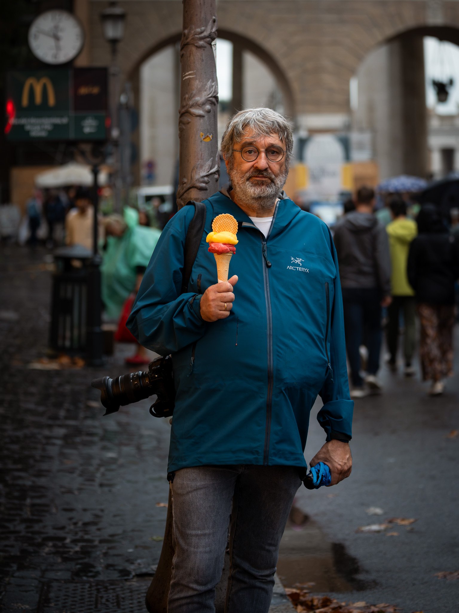 The photographer with gelato in the rain in Rome, Italy. Arc'teryx jacket, camera slung, holding an ice cream cone like it's the most natural thing. The McDonald's sign in the background is the detail that makes it.