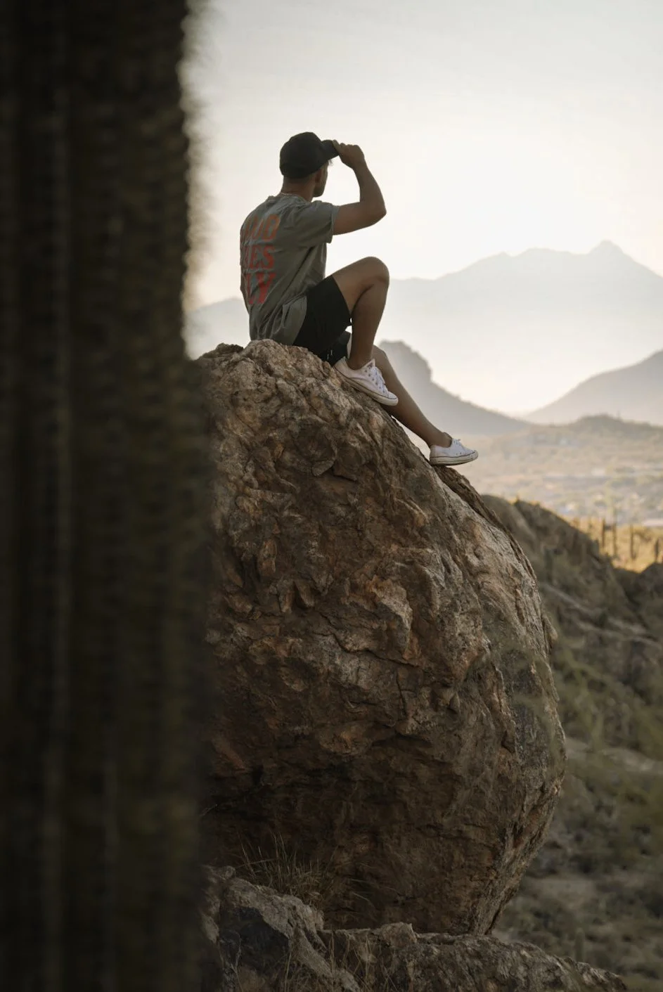 A self-portrait of Carlos Barrientos, sitting on a large rock, looking at mountains in the distance during sunset or sunrise. Photographed during a solo roadtrip in 2024 on a trailhead outside of Tucson, Arizona.