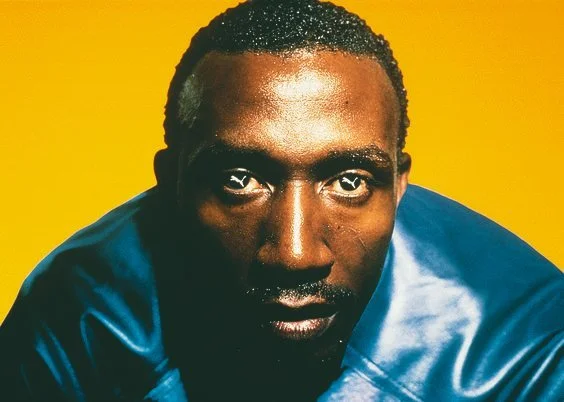 Close-up of a man with dark skin and short curly hair, wearing a shiny blue jacket, against a yellow background.
